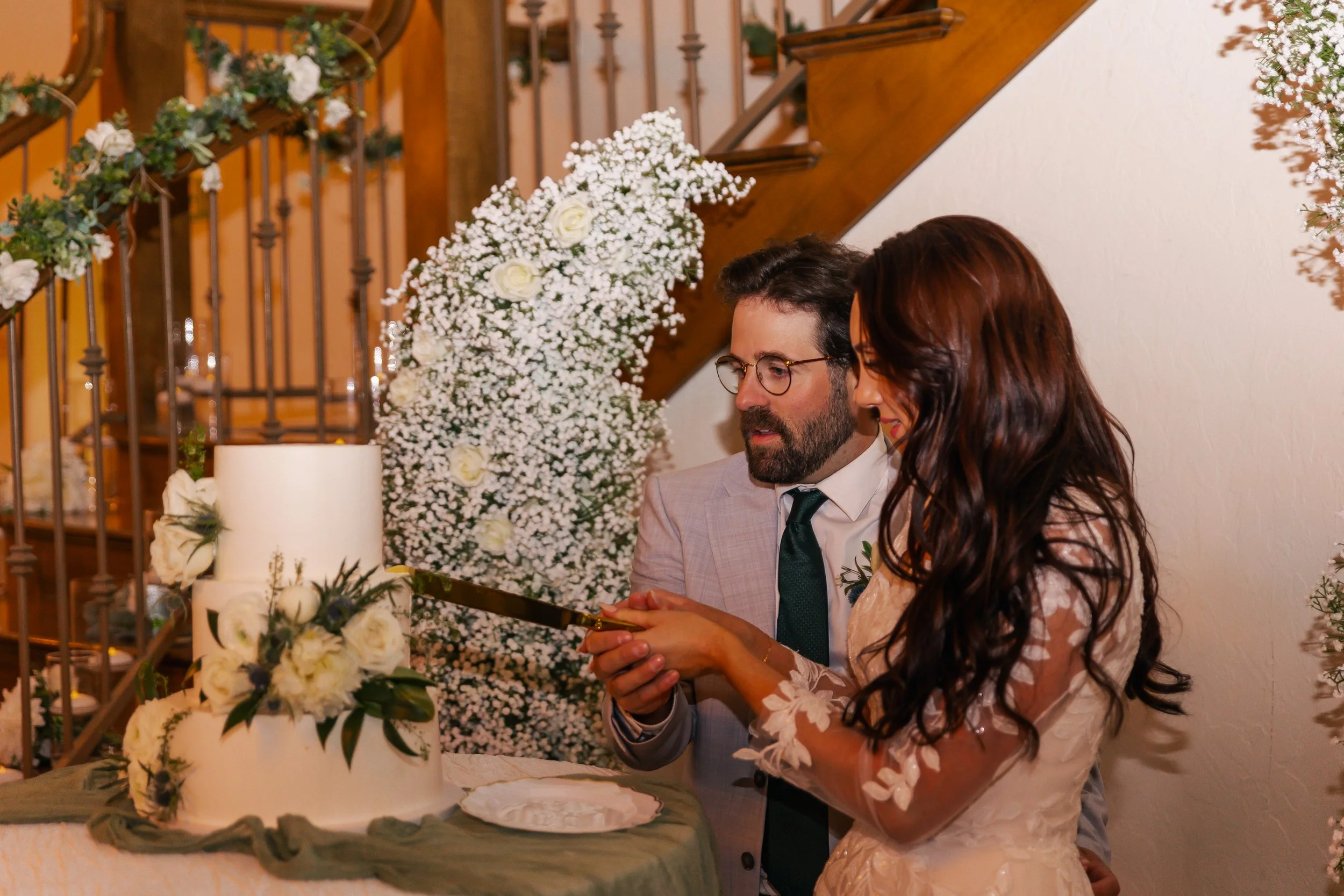 A couple cutting a wedding cake together at a celebration indoors, decorated with white flowers and greenery.