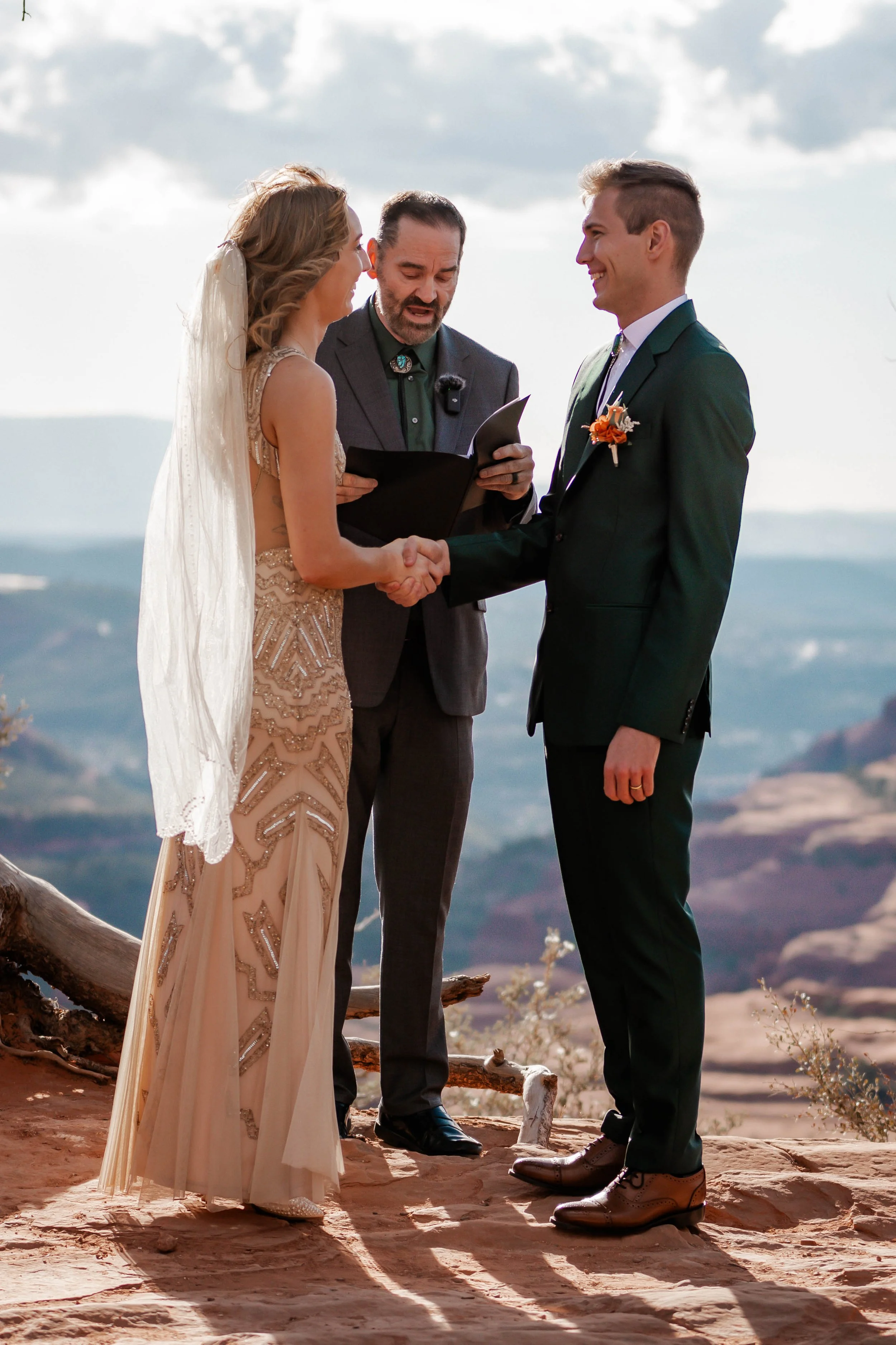A couple gets married outdoors, holding hands and smiling at each other while shaking hands. An officiant reads from a book, with a mountain landscape in the background.