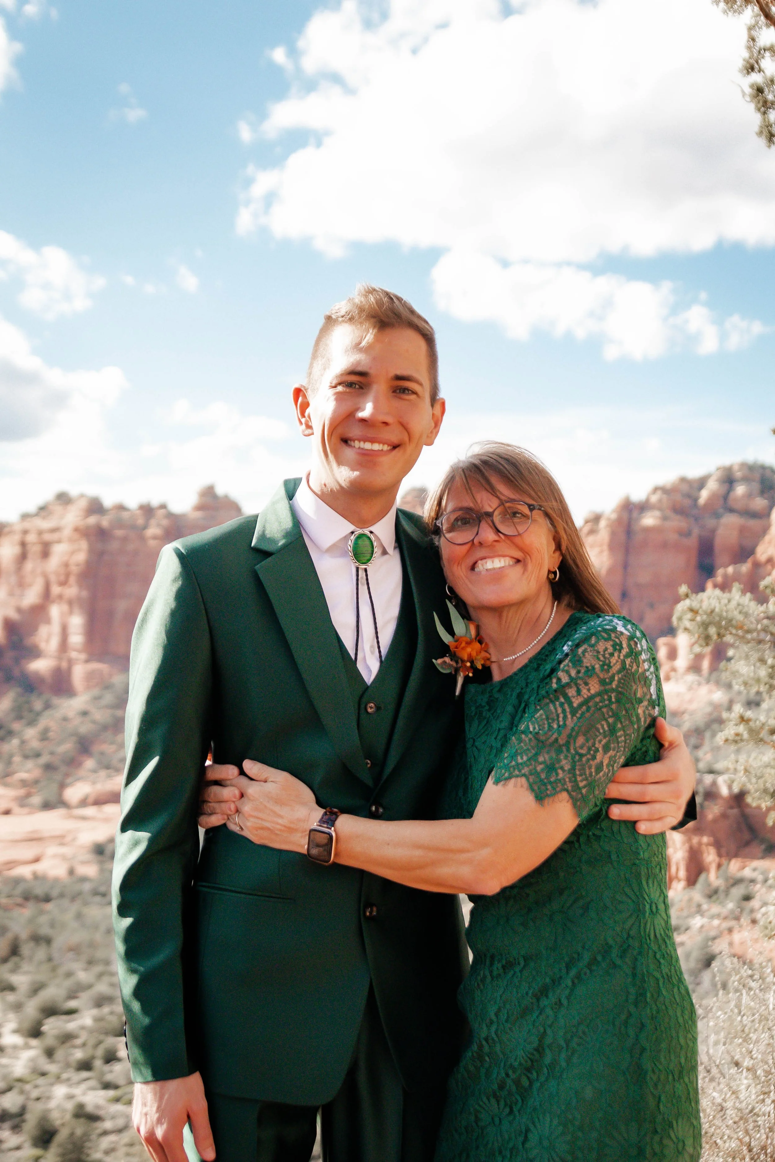 A young man in a green suit and an older woman in a lace green dress embrace and smile outdoors with desert mountains and a blue sky with white clouds in the background.