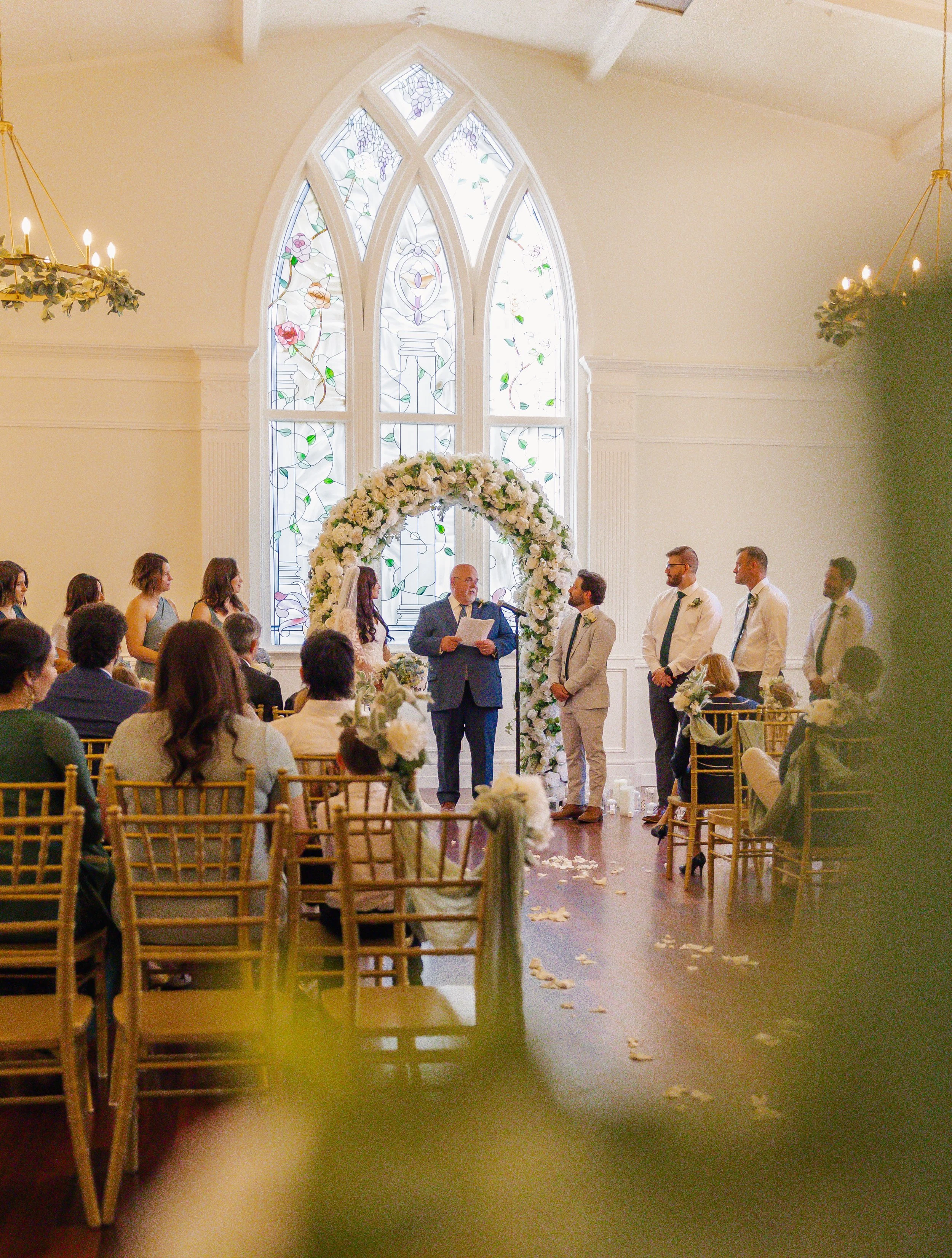 A wedding ceremony inside a church with a tall, stained glass window in the background. The officiant, a man in a suit, is reading from a paper while the bride and groom stand before him. The bride wears a white wedding dress, and the groom wears a l
