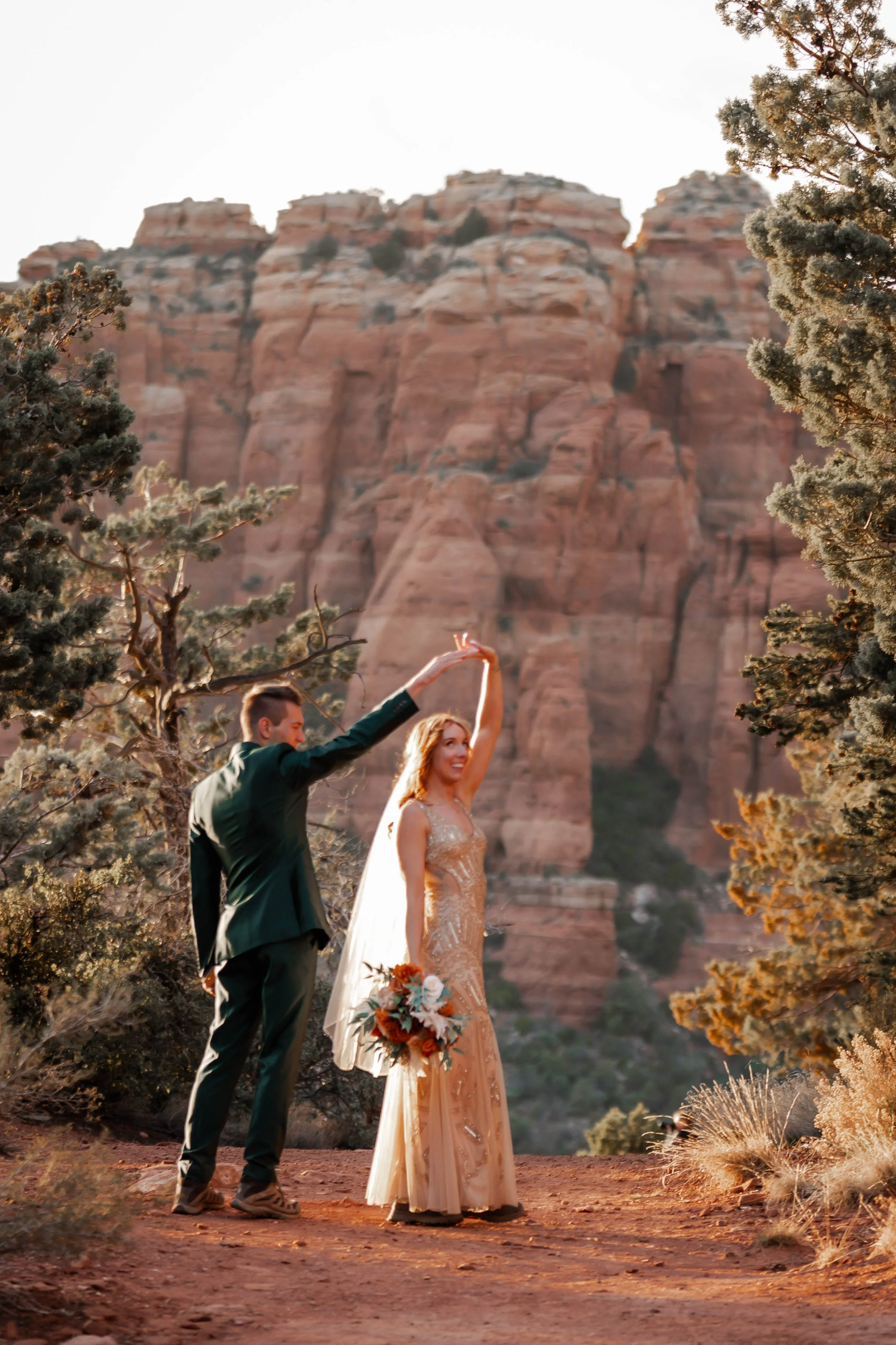 A couple dancing outdoors at sunset with a red rock canyon in the background, one holding a bouquet of flowers.