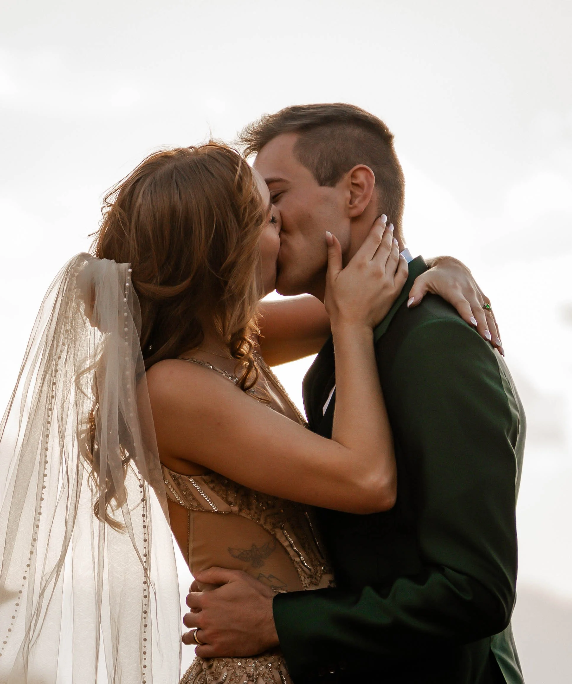 A couple sharing a kiss outdoors on their wedding day, with the bride wearing a veil and the groom in a dark suit.