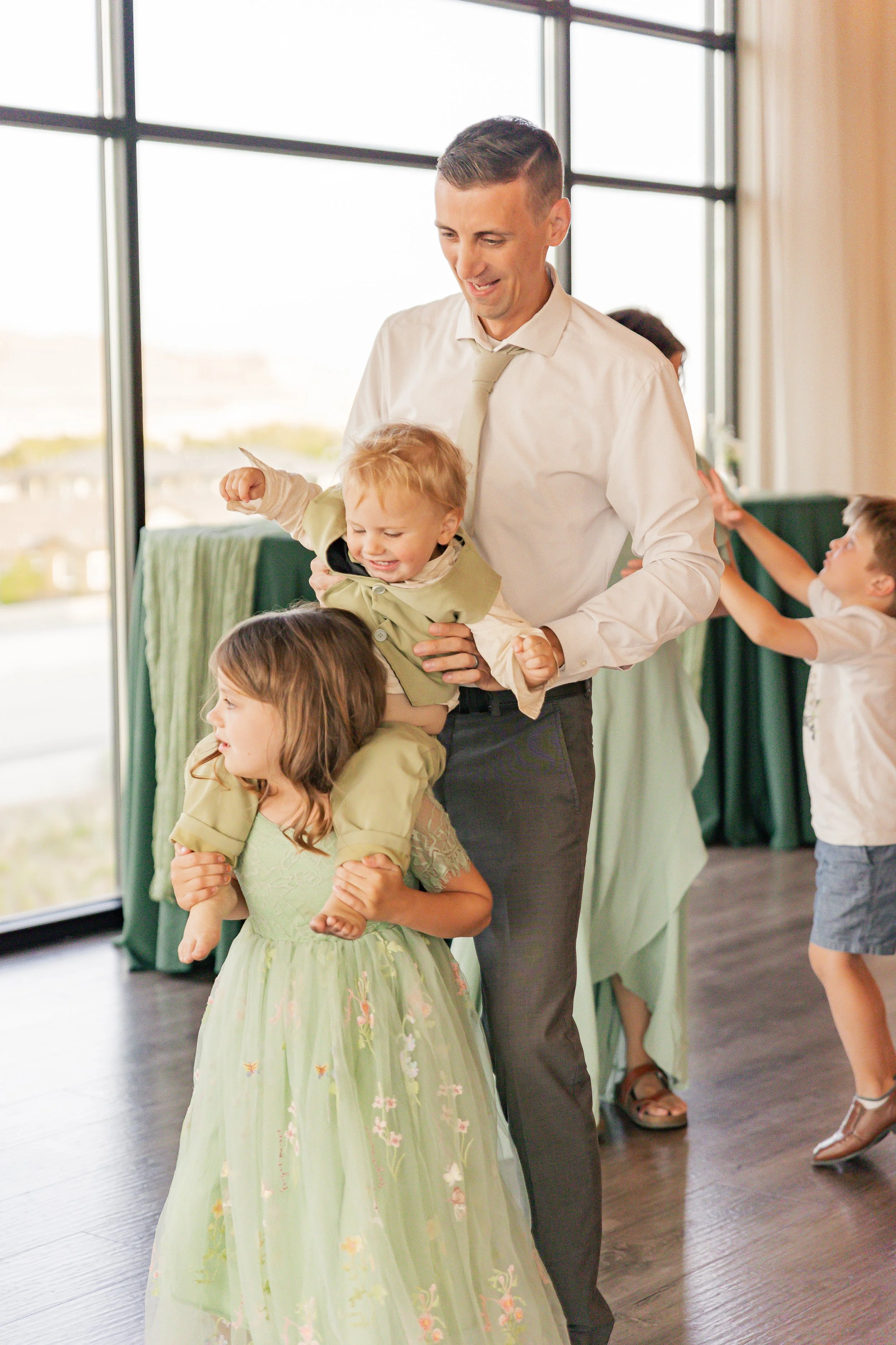 A man is holding two children, one on his shoulders and one in his arms, as they dance or play indoors near large windows with a view outside. Other children are visible in the background, also engaged in playful activities.
