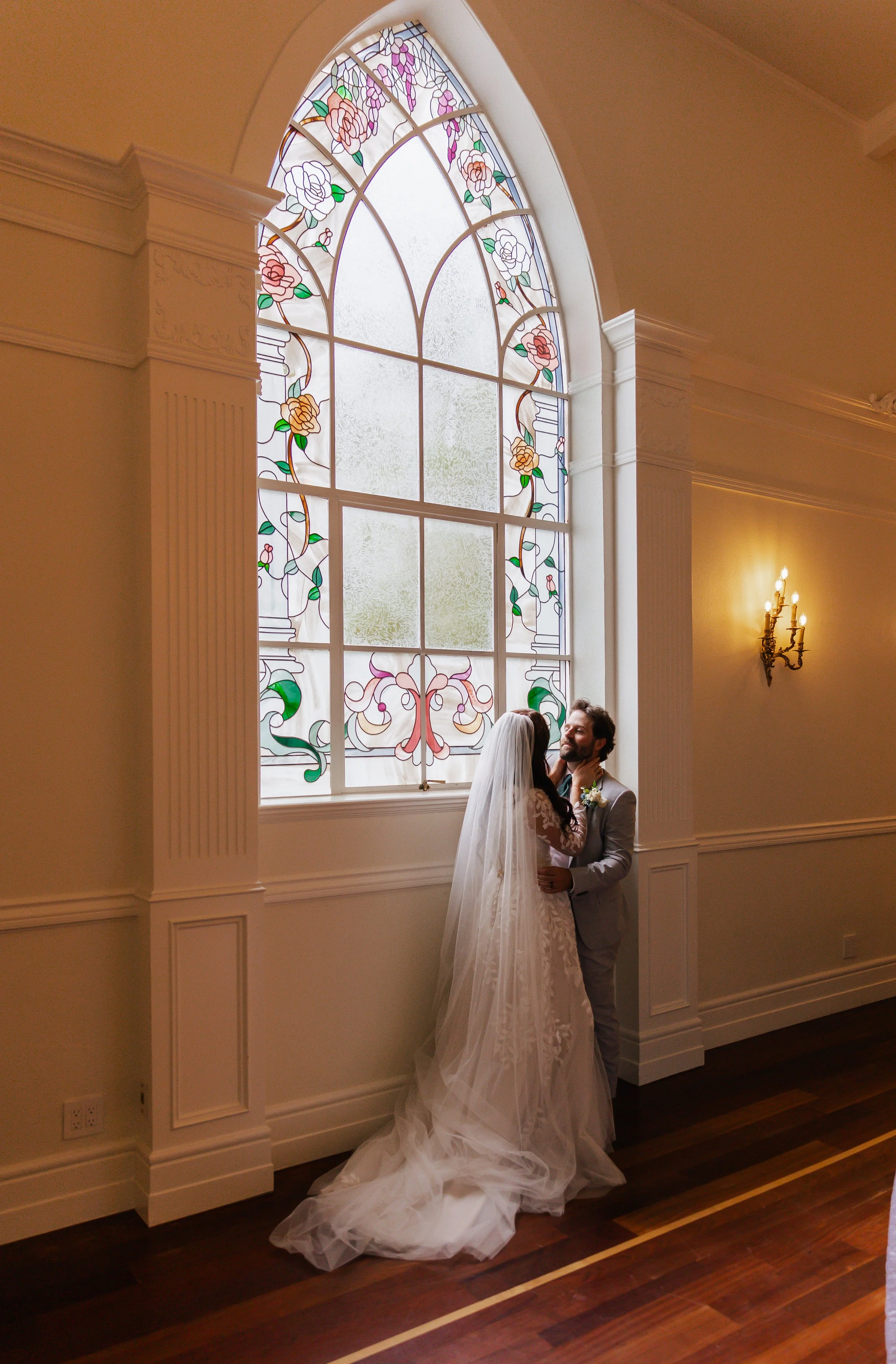 Bride and groom sharing a kiss standing by a large stained glass window in a wedding venue.