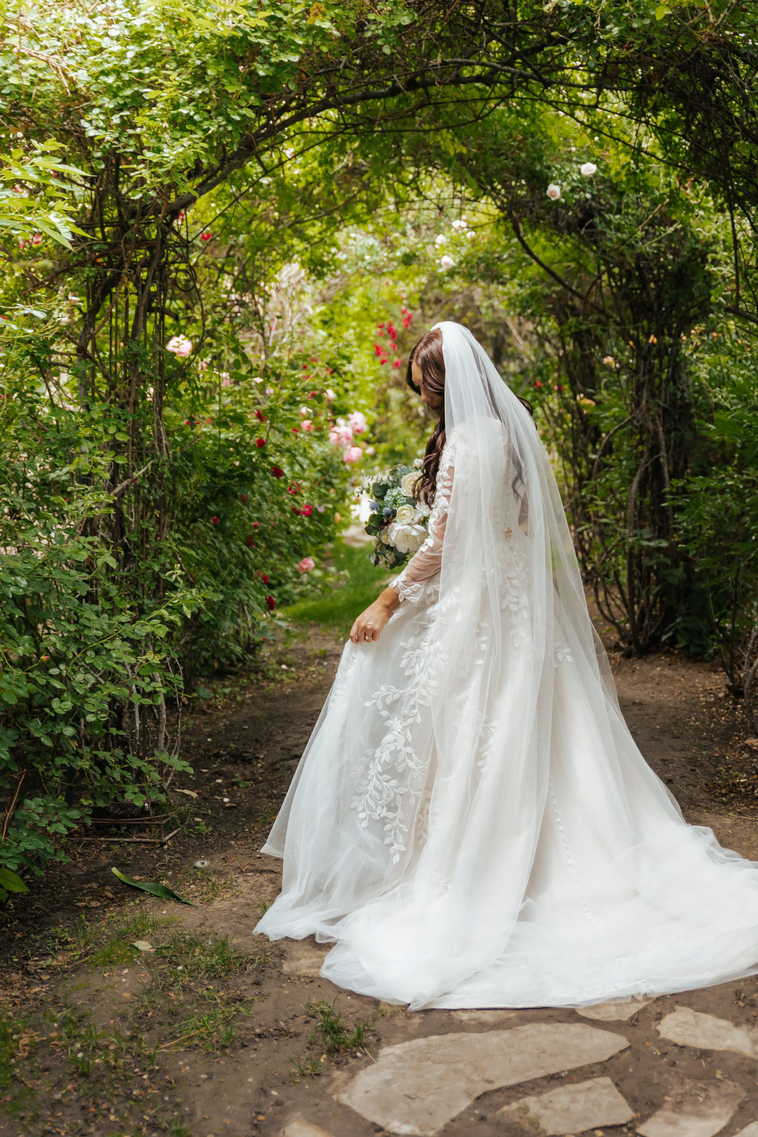 A bride in a white wedding gown and veil holding a bouquet of white roses, standing under a green archway of flowering bushes on a garden path.