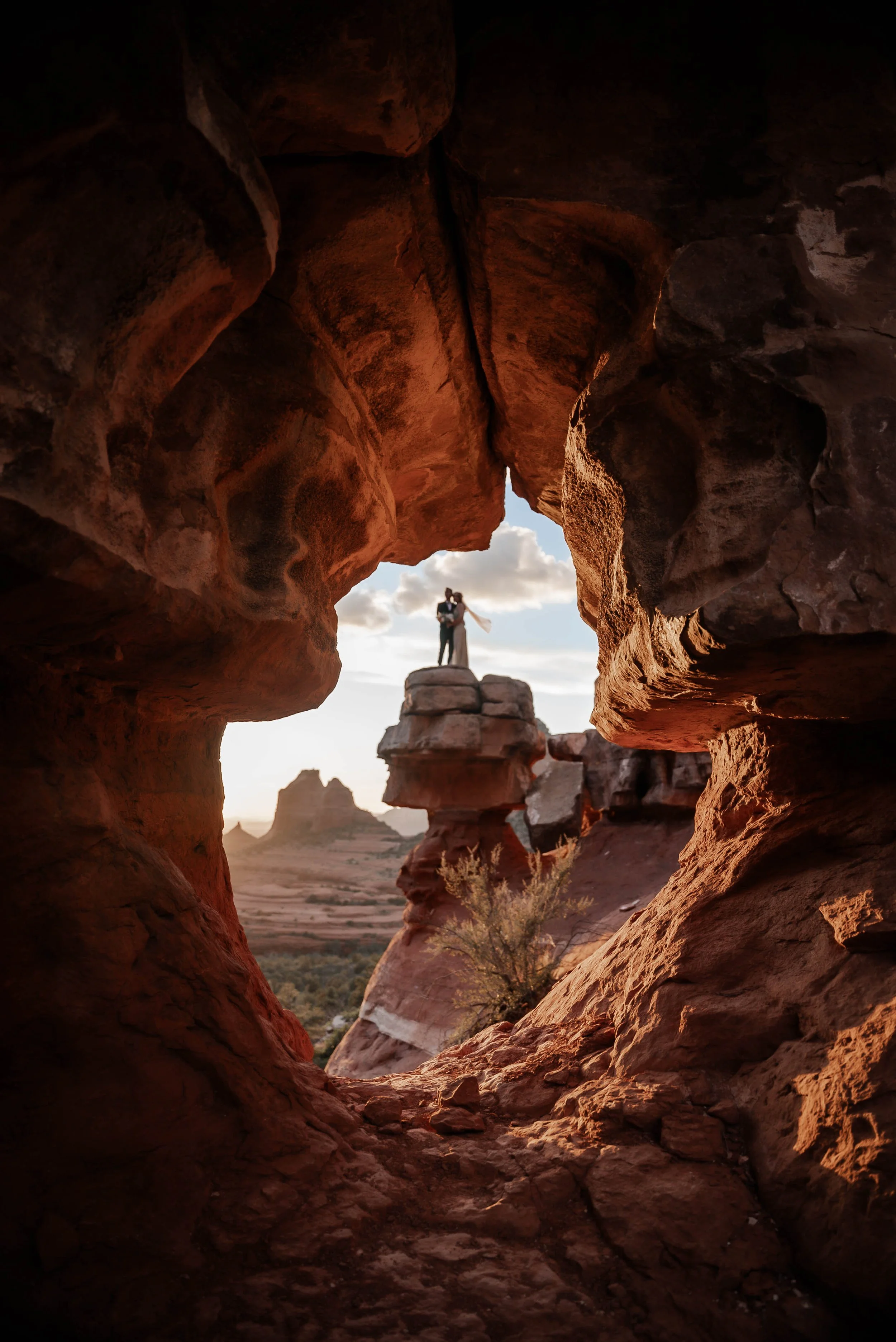 A couple standing on a rock formation in a desert landscape, framed by a natural rock window with reddish-brown rocks and a cloudy sky.