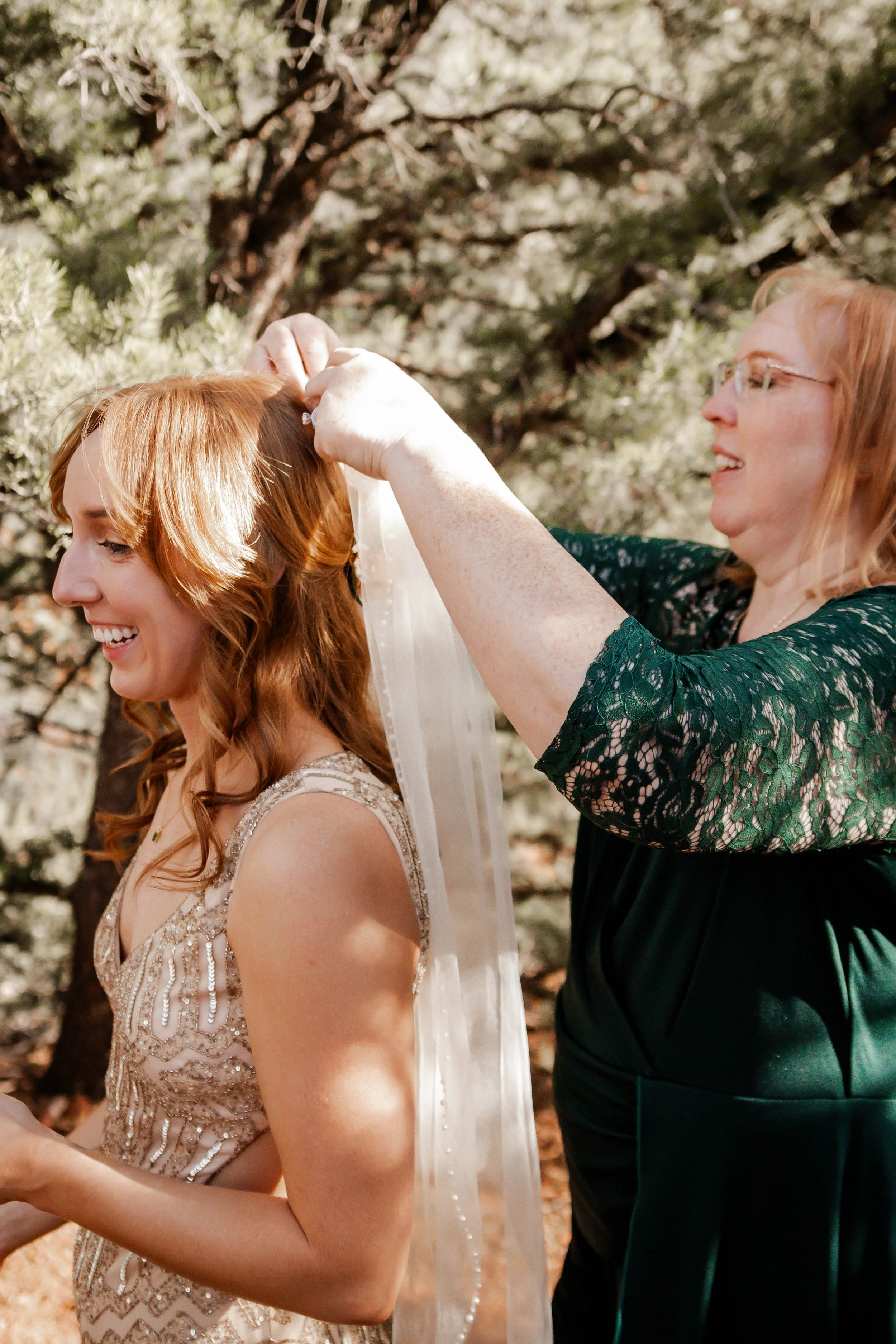 A woman helping a bride put on a veil outdoors during daytime, with trees in the background.