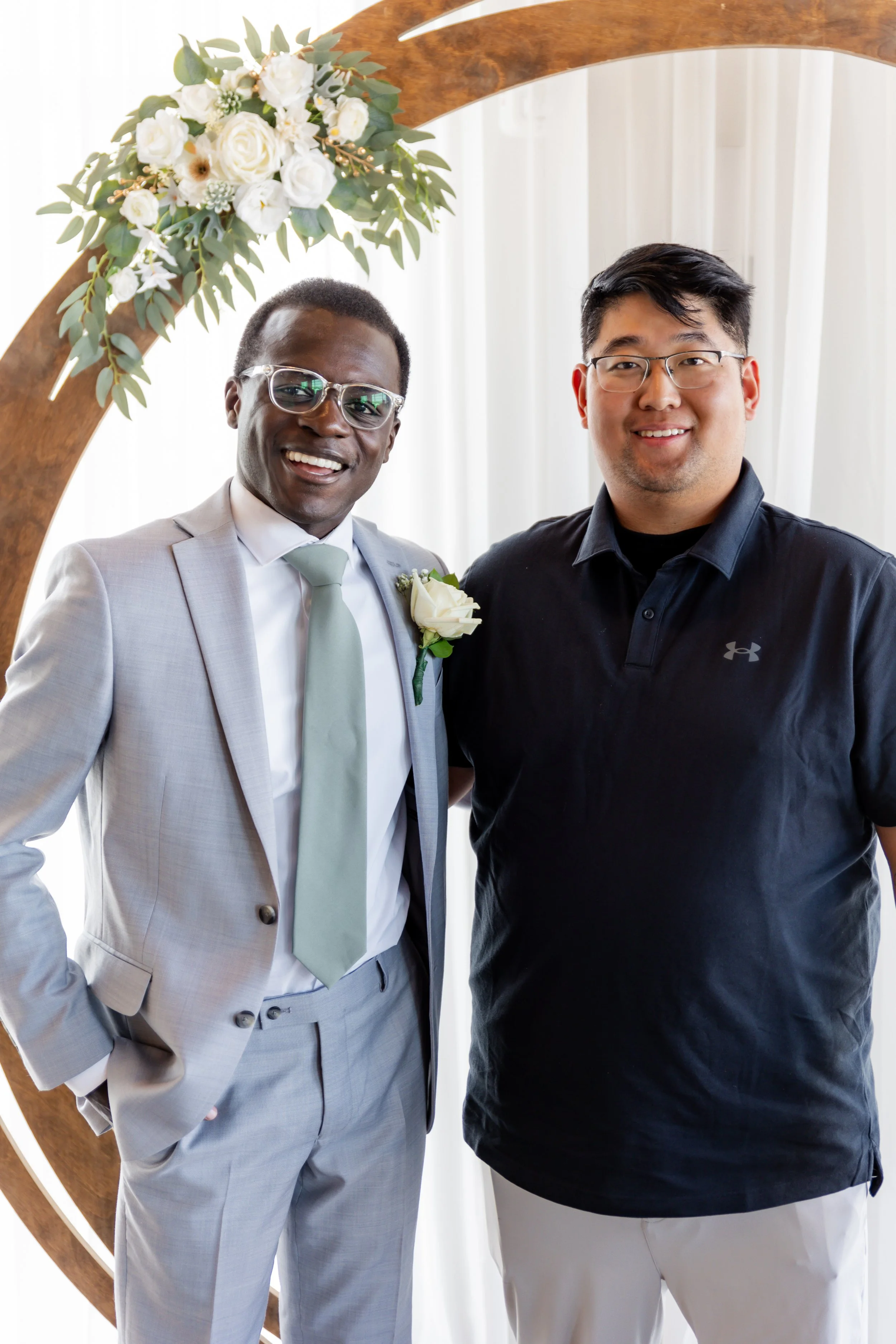 Two men standing in front of a floral wedding arch, smiling at the camera. One is dressed in a light gray suit with a white shirt and light gray tie, wearing glasses. The other is dressed in casual black Under Armour polo and white pants, wearing gla