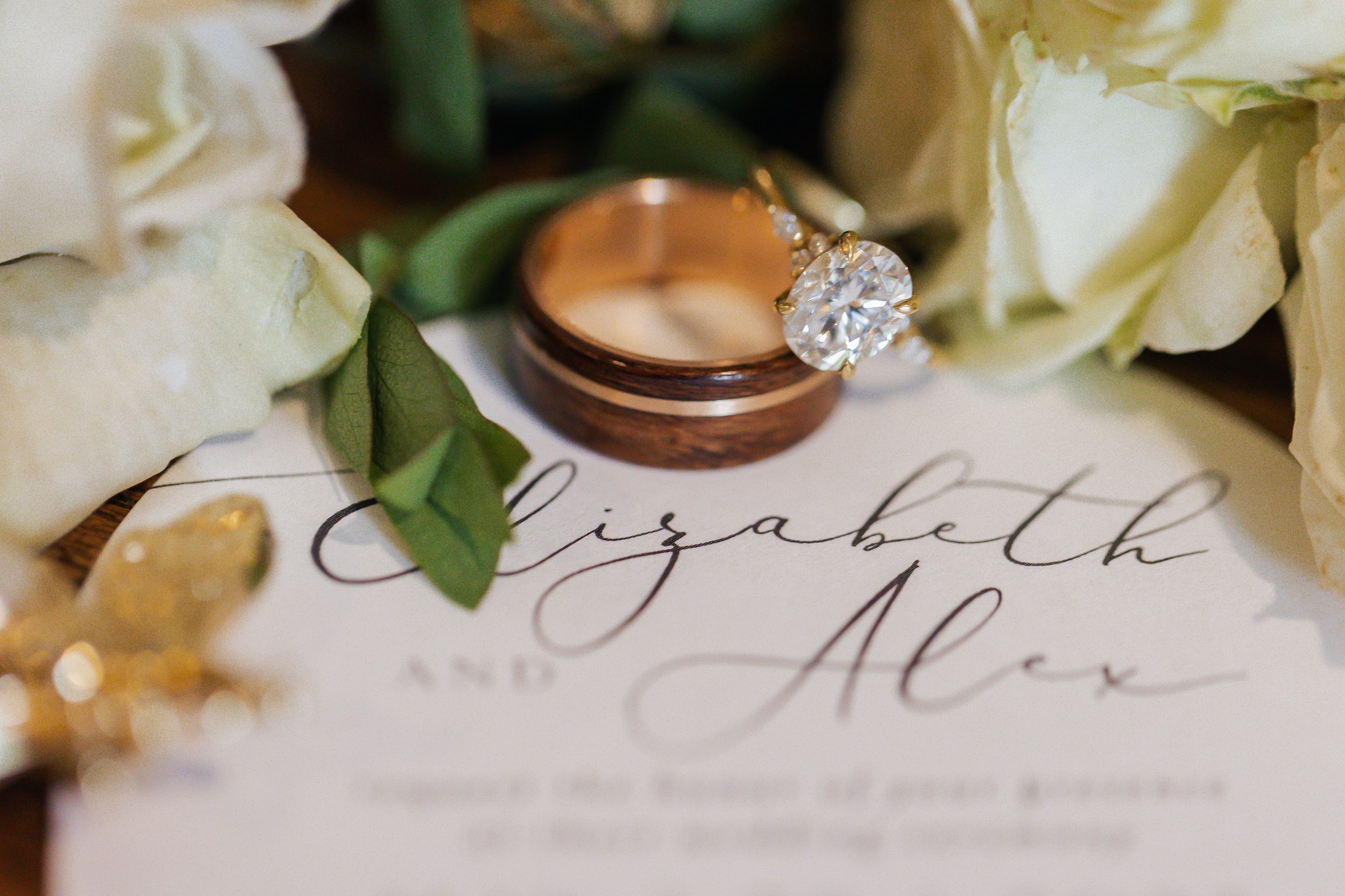 Close-up of a diamond engagement ring and a wedding band on a wedding invitation that reads 'Elizabeth and Alex,' surrounded by white roses and green leaves.