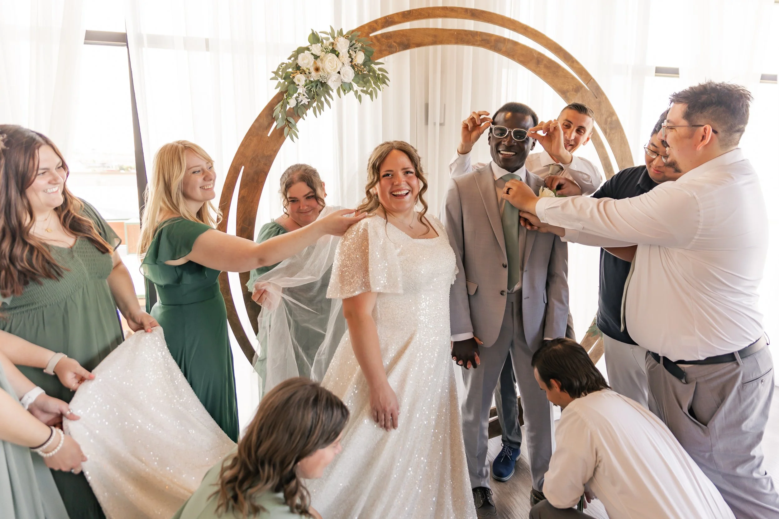Bride and groom with bridal party at wedding, standing in front of a circular floral decoration, people smiling and helping the bride with her veil.