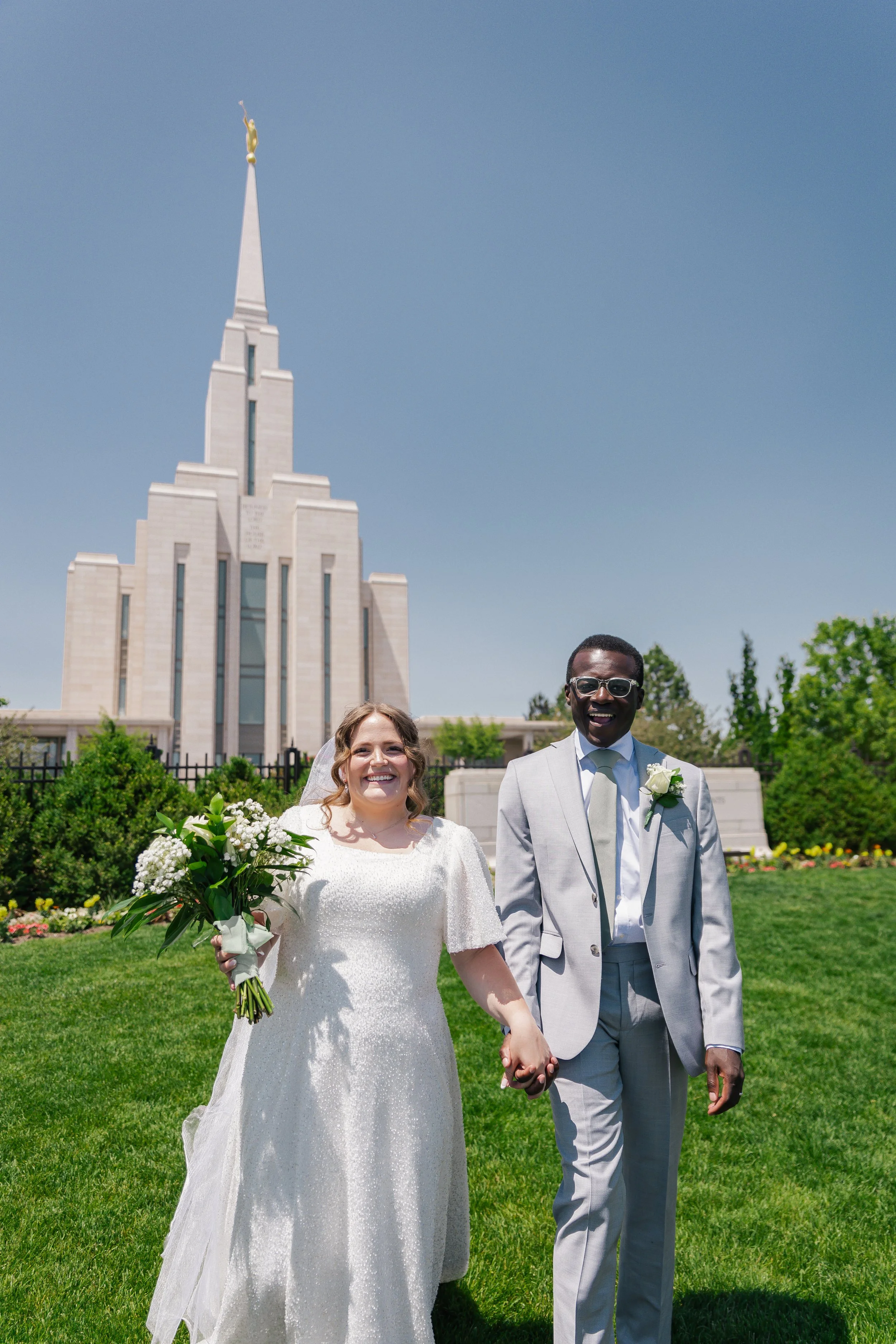 A happy bride and groom holding hands and walking on a grassy lawn, with oquirrh mountain temple in the background.