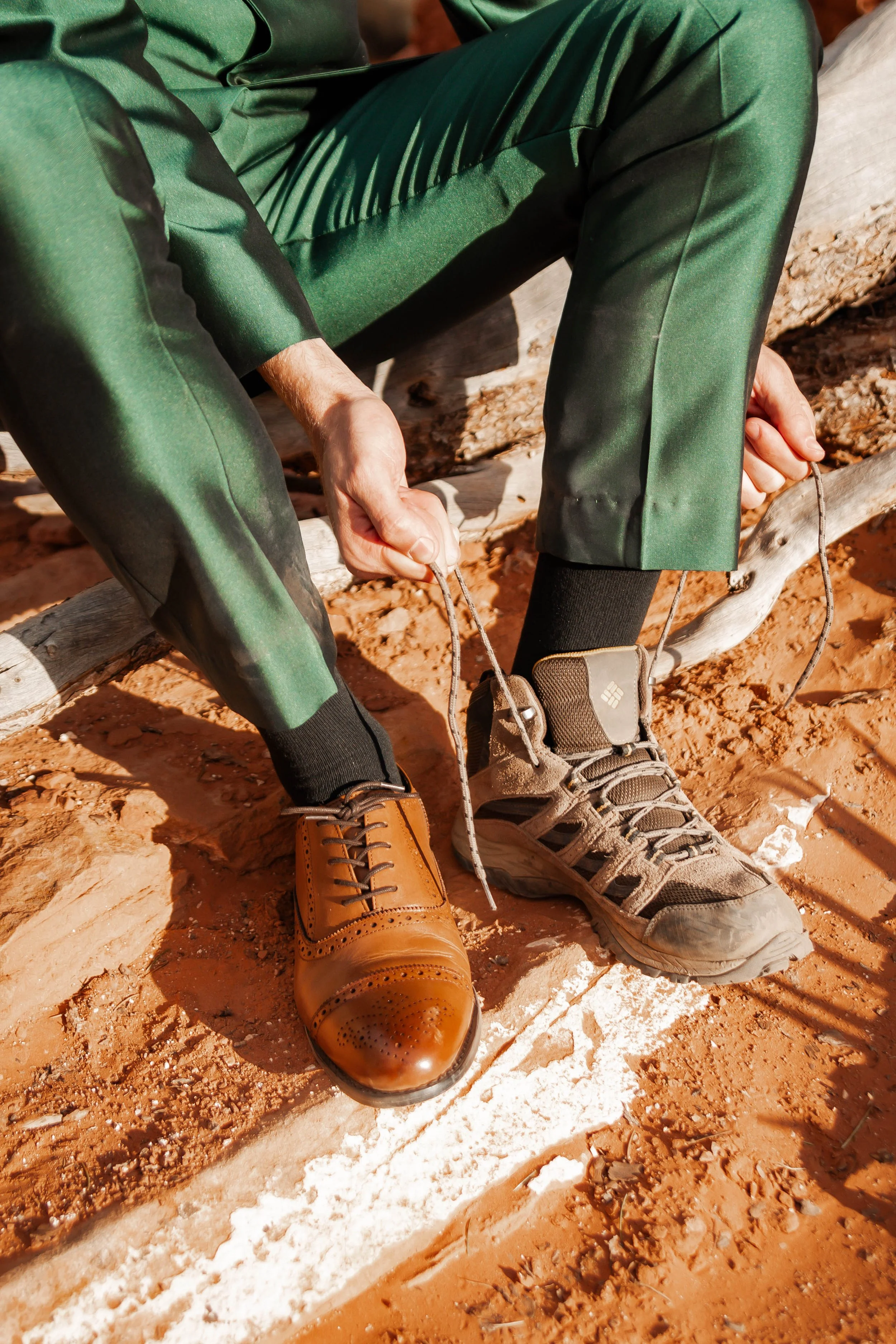 Person wearing one polished brown dress shoe and one rugged hiking boot, tying their shoelaces on a red dirt ground.