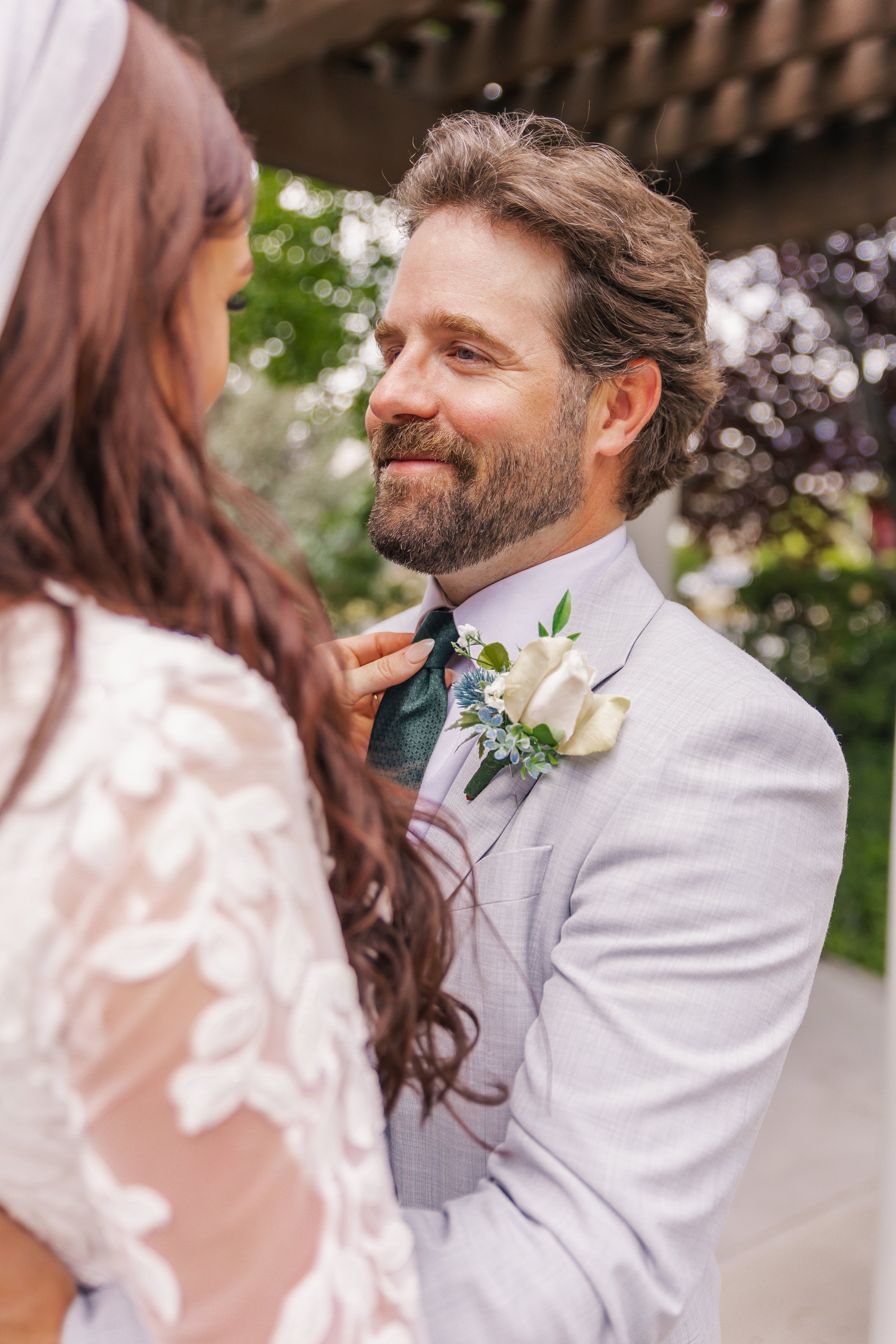 A bride and groom at their wedding, with the bride adjusting the groom's tie. The groom is wearing a light gray suit with a white shirt, black tie, and a boutonniere with white flowers. The bride has long, wavy hair and is wearing a white lace dress.