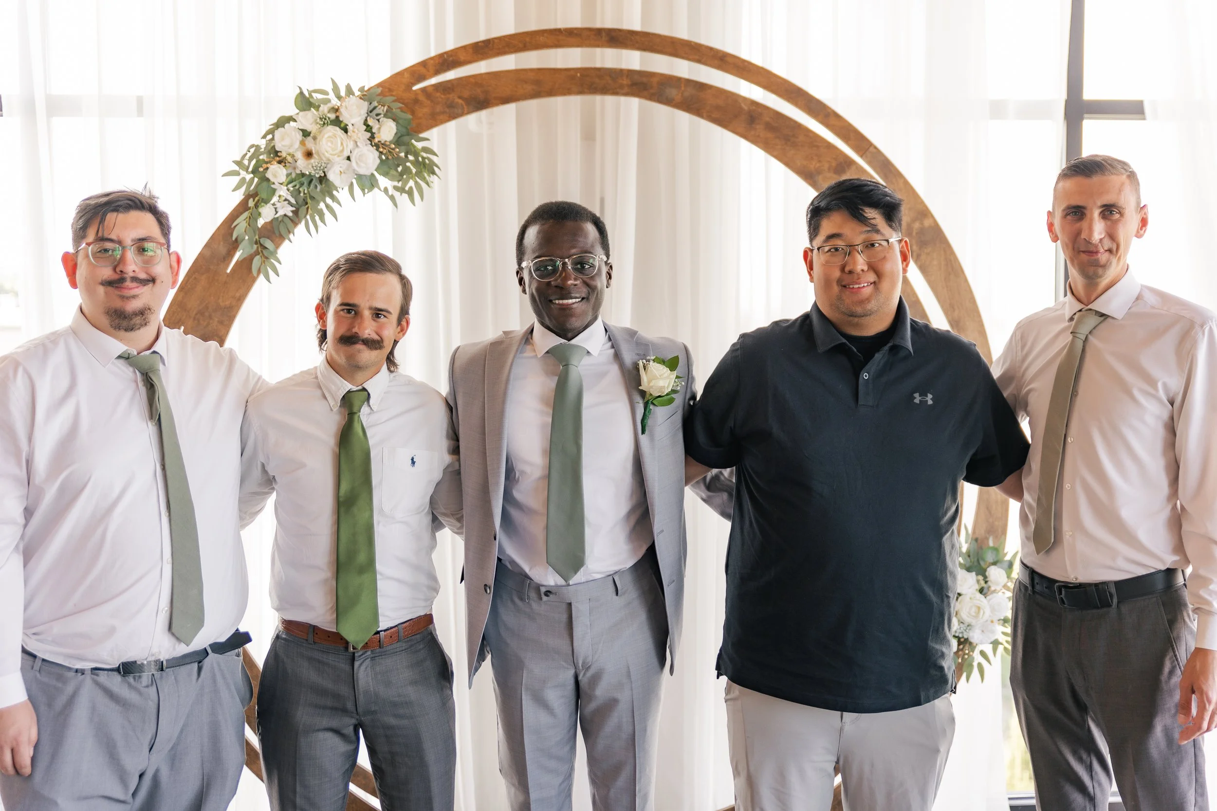 Group of six men standing in front of a wedding arch with white flowers, smiling.