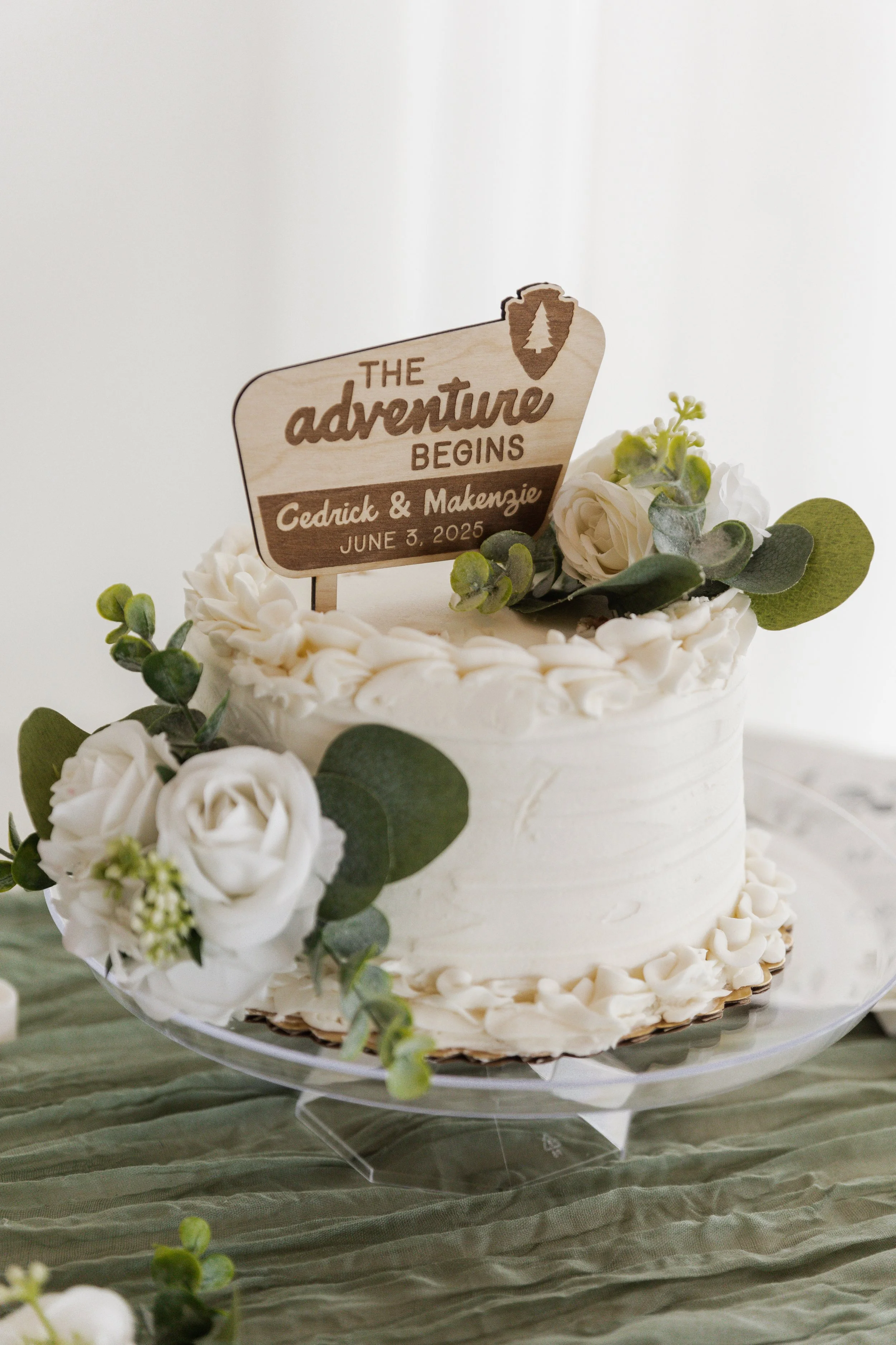A white wedding cake decorated with white flowers and greenery, topped with a wooden sign that reads 'The adventure begins, Cedrick & Makenzie, June 3, 2025'.