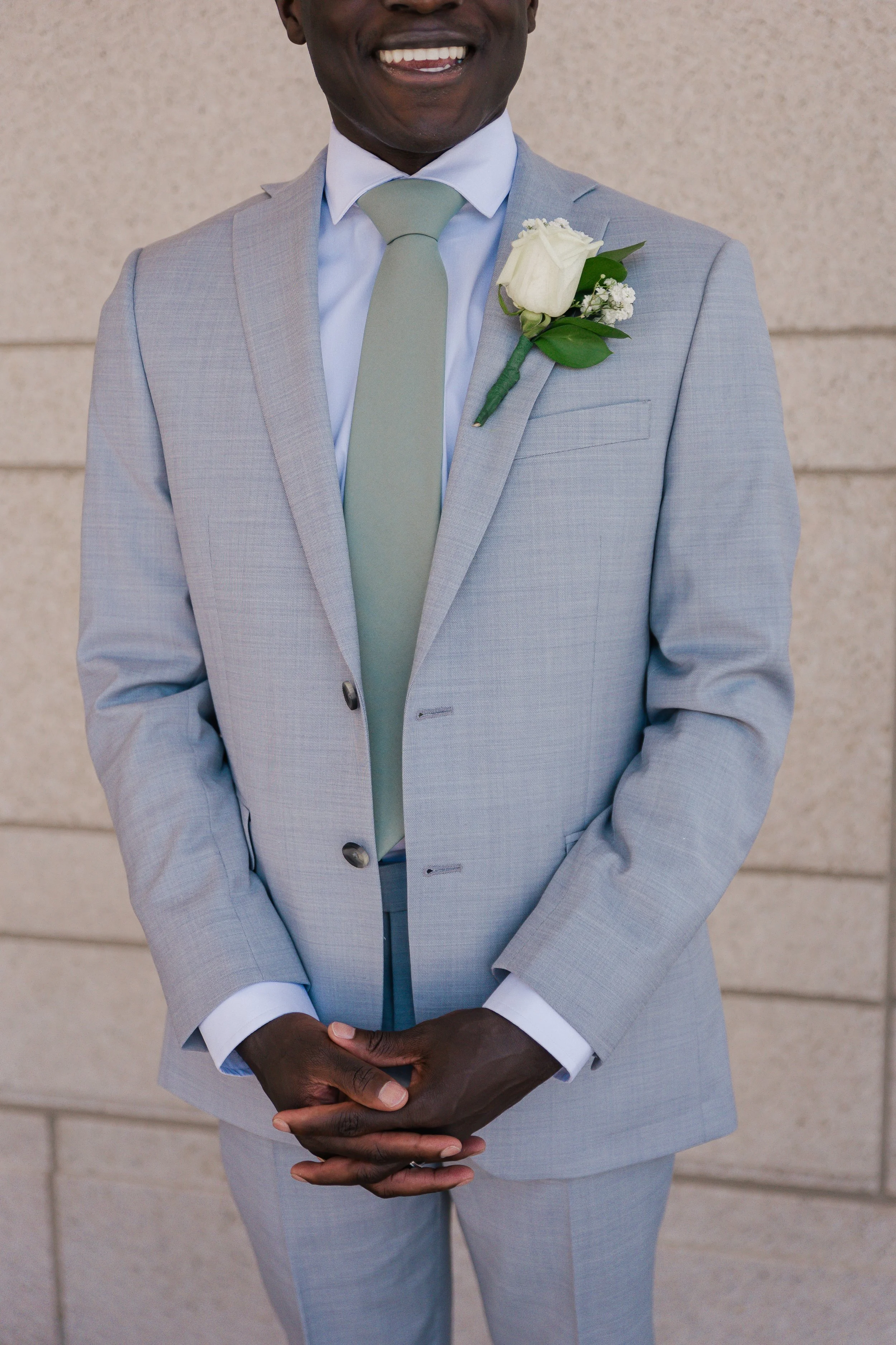 A man in a light gray suit with a white shirt and pale green tie, smiling with hands clasped in front, standing against a beige wall, with a white rose boutonniere pinned to his lapel.