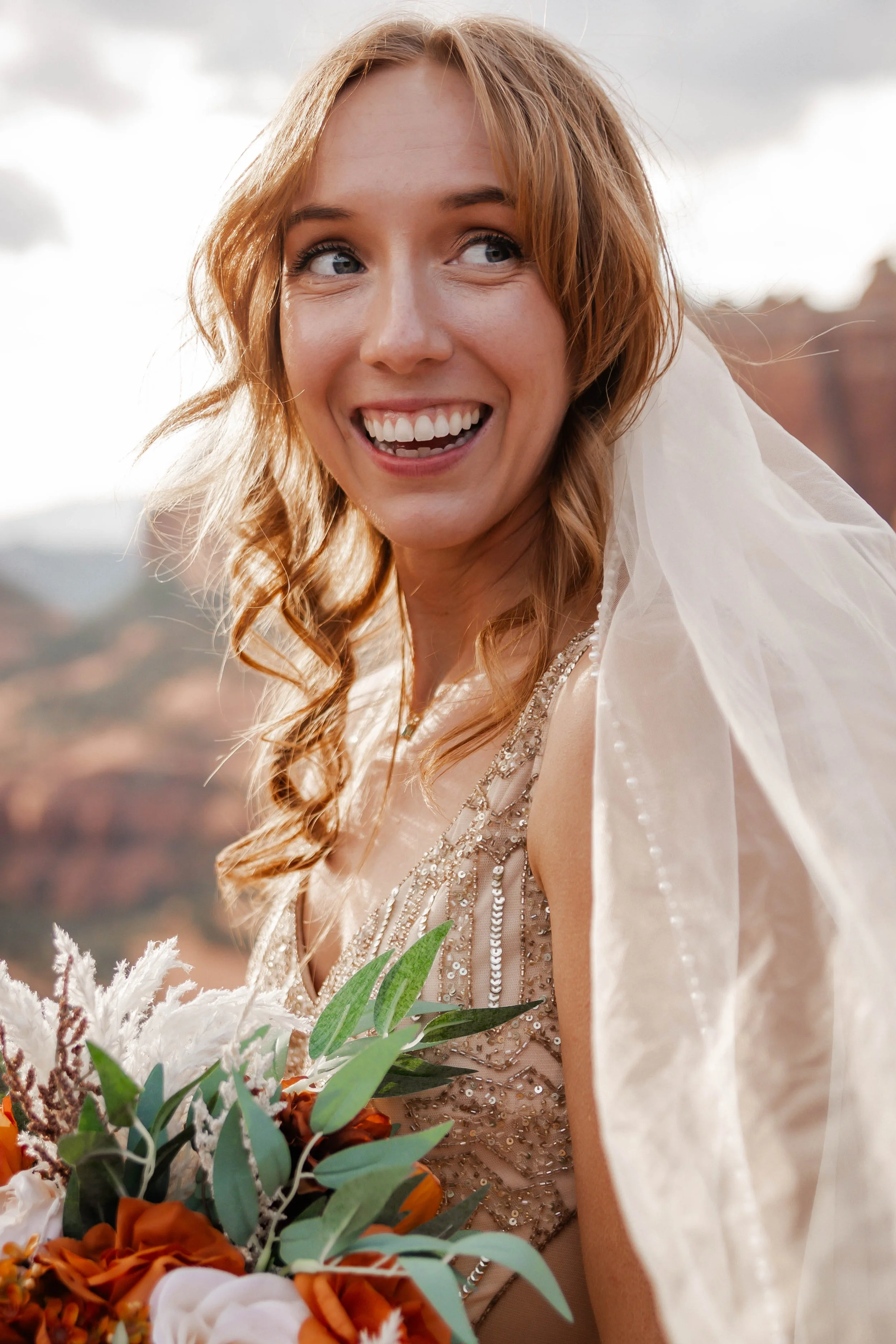 A smiling woman with red hair and blue eyes holding a bouquet, standing outdoors with mountains in the background.