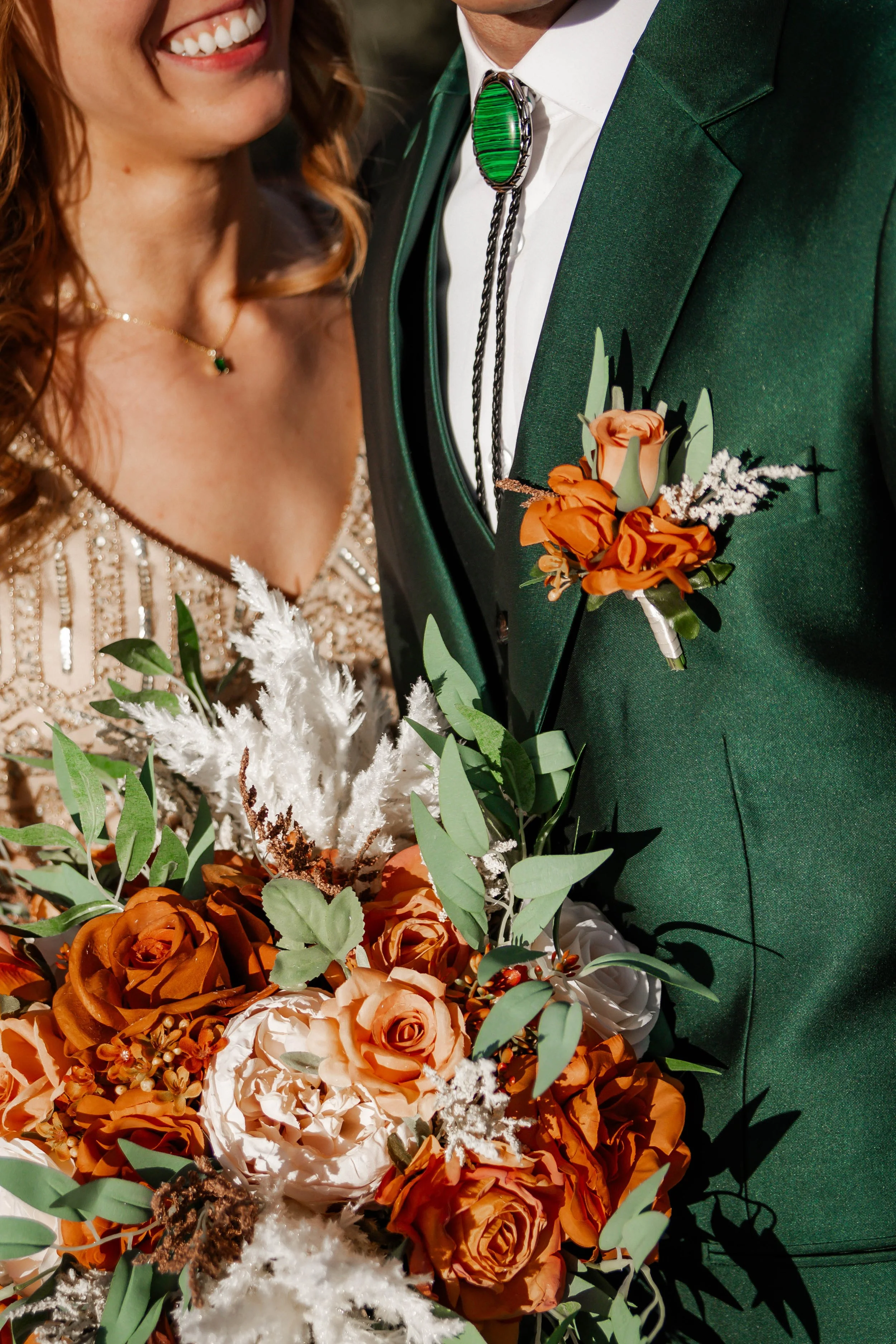 Close-up of a bride and groom, showing only their torsos, holding a bouquet of orange and white roses with greenery. The groom is wearing a green suit with a rose boutonniere and a bolo tie, while the bride is wearing a beaded dress with a gold neckl
