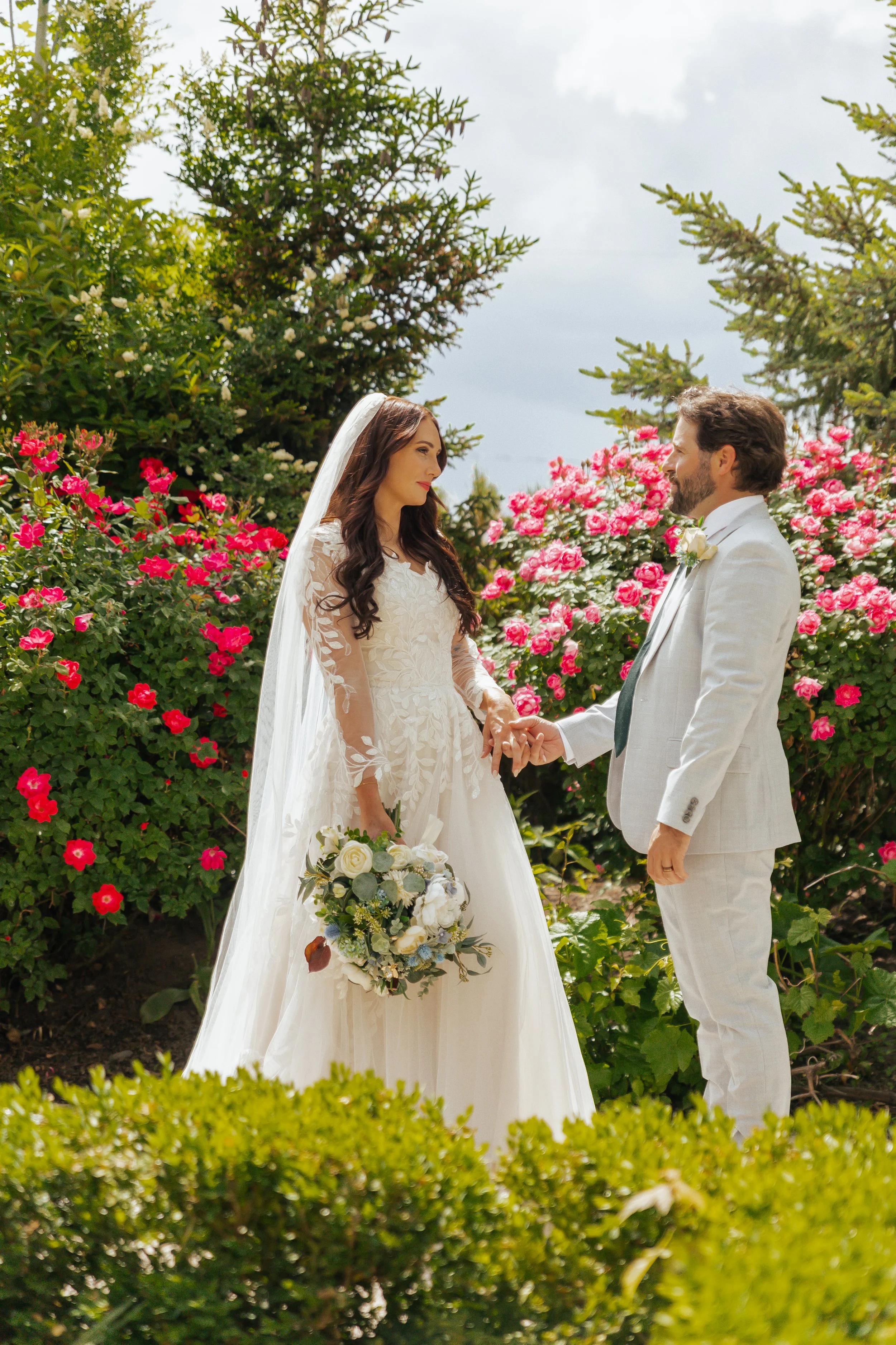 A bride and groom holding hands during their wedding in a garden with pink and red flowers.