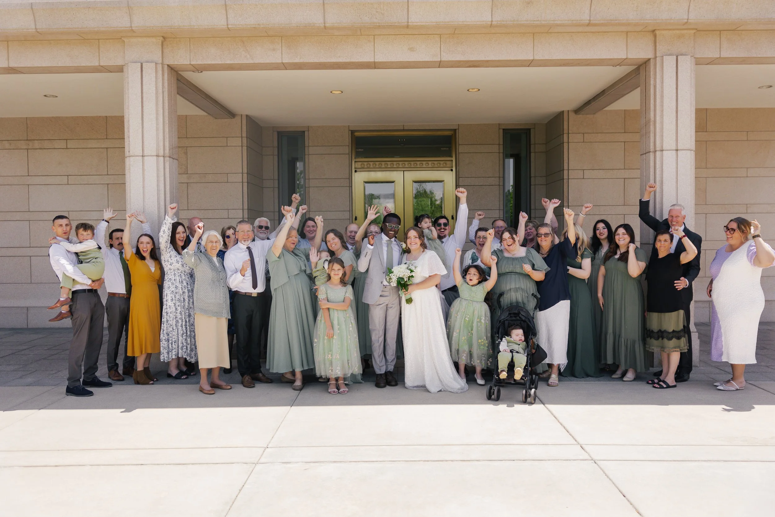 A large group of people gathered in front of a building, celebrating a wedding, with many raising their fists in the air, including a bride and groom at the center.