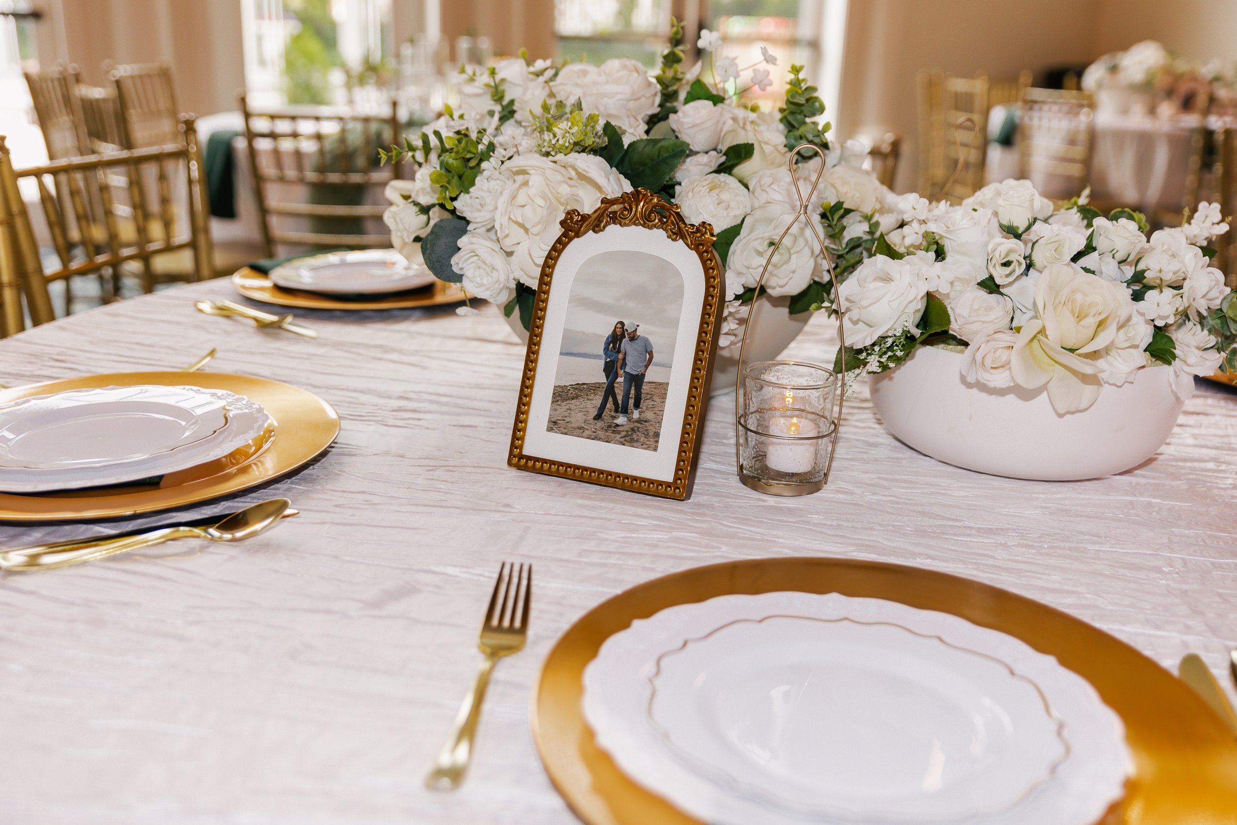 A table decorated with a large floral centerpiece of white flowers, gold chargers, white plates, gold cutlery, a photo frame with a picture of a couple, and candles, set in a well-lit room with chandelier and window in the background.