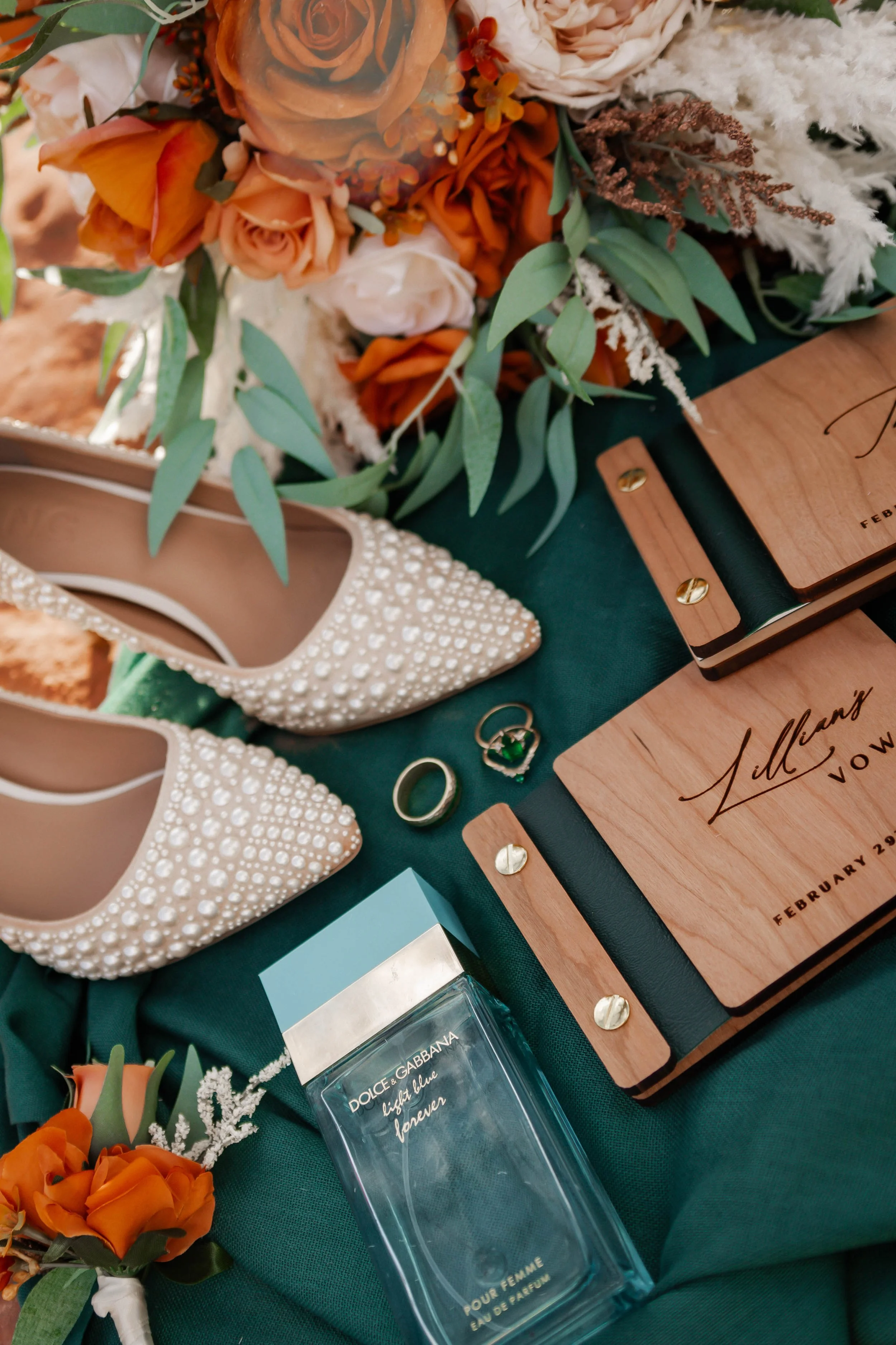 Wedding or special event display with white pearl-embellished high heels, orange and cream flowers, greenery, small wooden signs, a bottle of Dolce & Gabbana perfume, a ring with a green gemstone, and a small floral boutonniere.