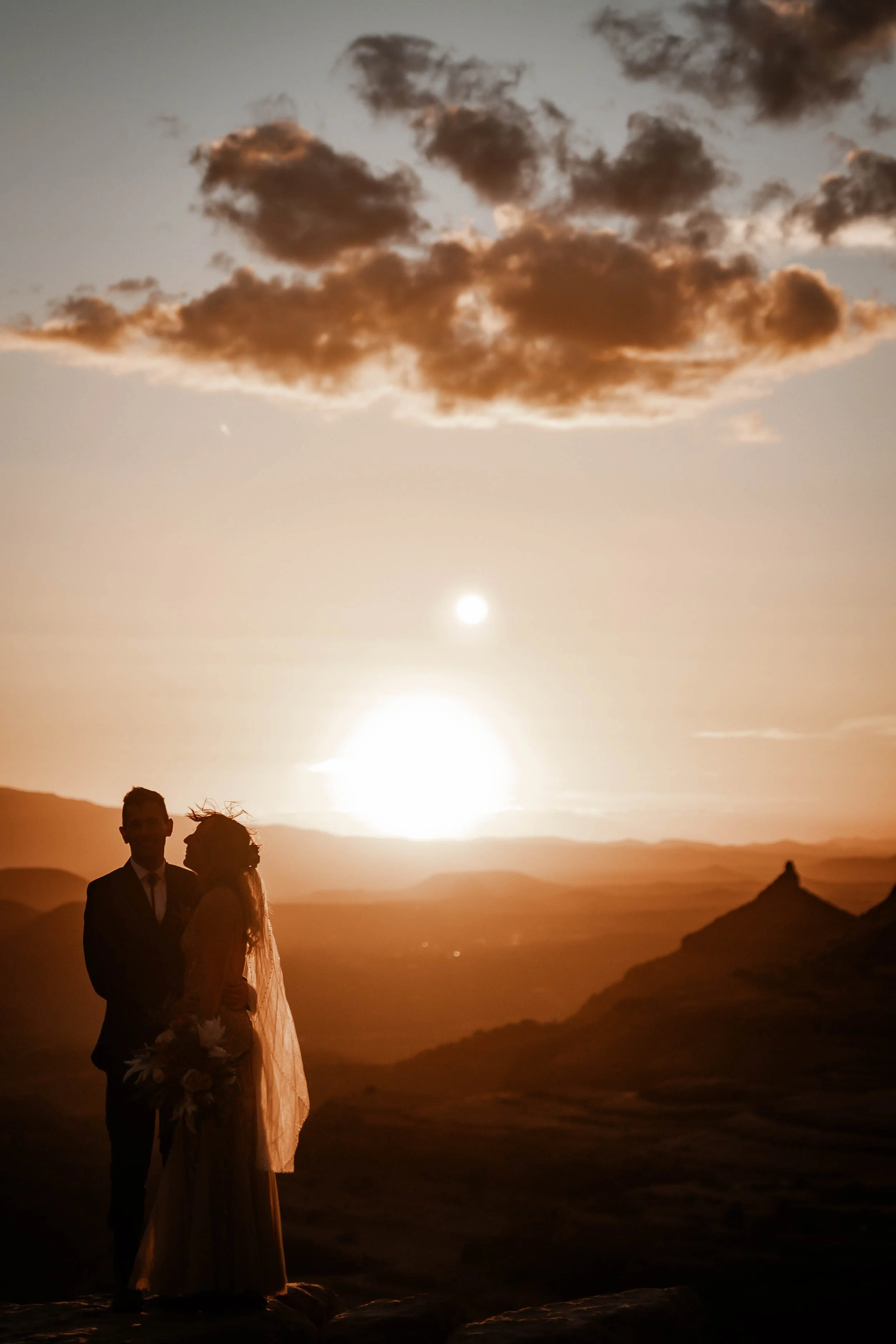 Silhouette of a bride and groom at sunset in a mountainous landscape with a large sun and scattered clouds.