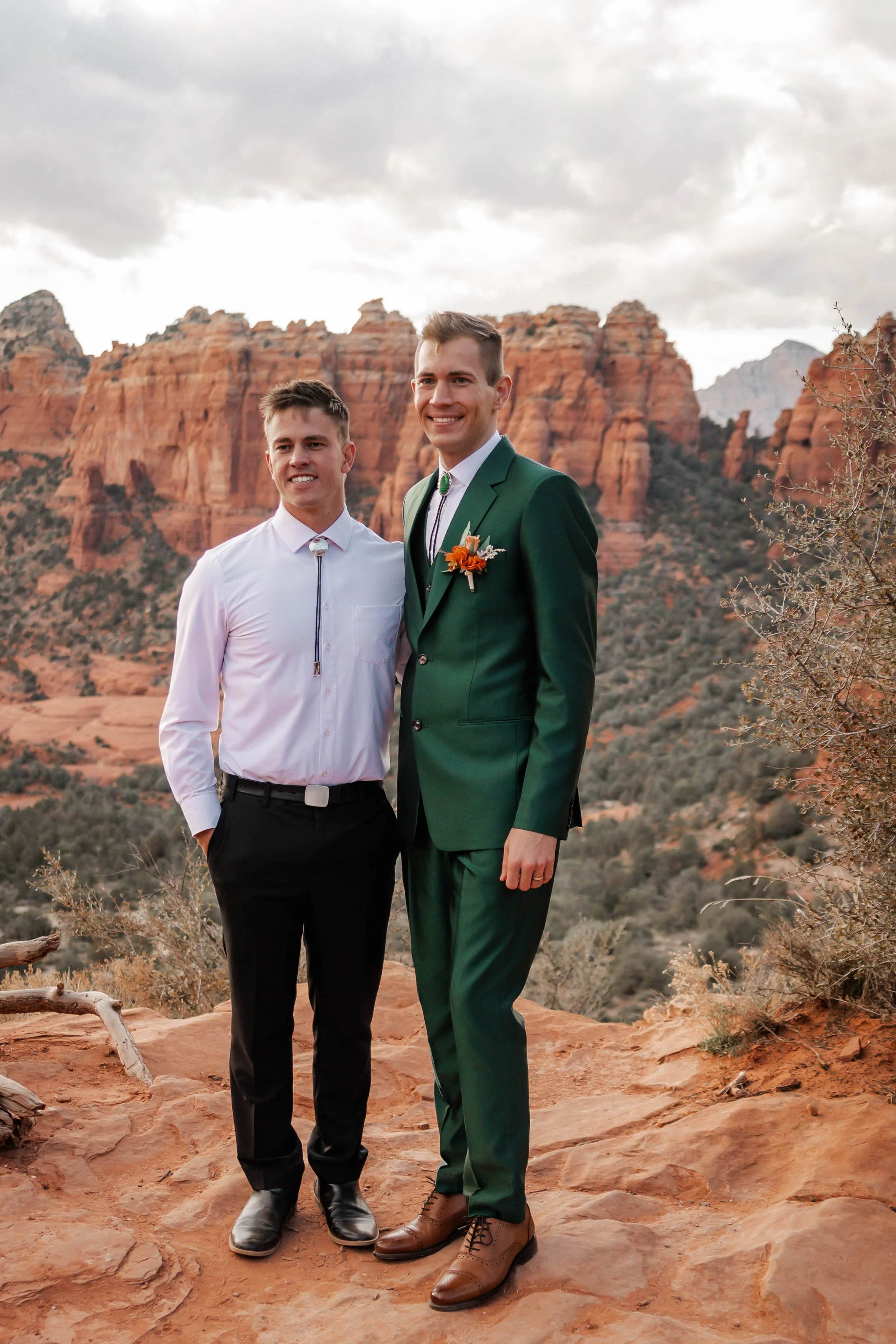 Two men standing on rocky terrain in front of red rock formations, one in a white shirt and black pants, the other in a green suit with a boutonnière, during cloudy weather.