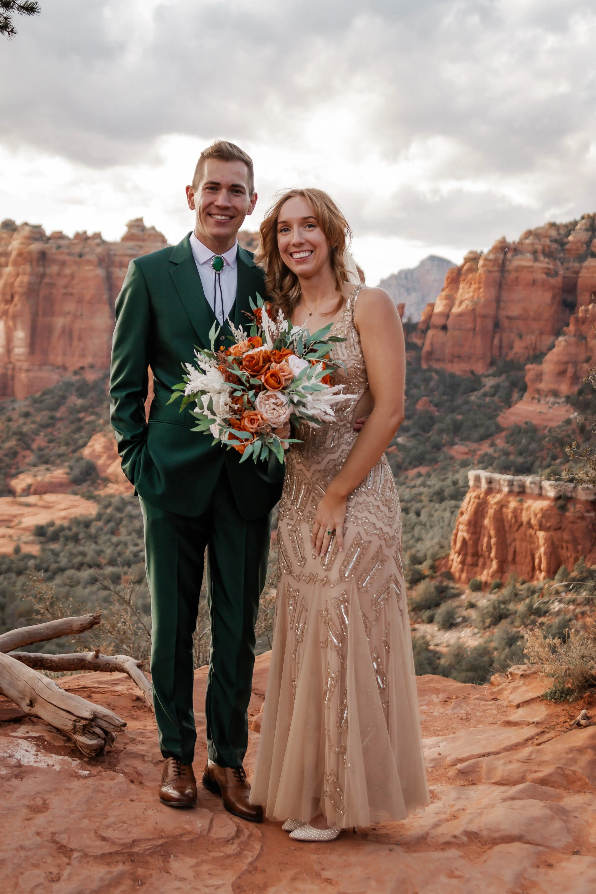 A wedding couple standing on a rocky ledge of a canyon, smiling, with the groom in a green suit holding a bouquet and the bride in an embellished gown.
