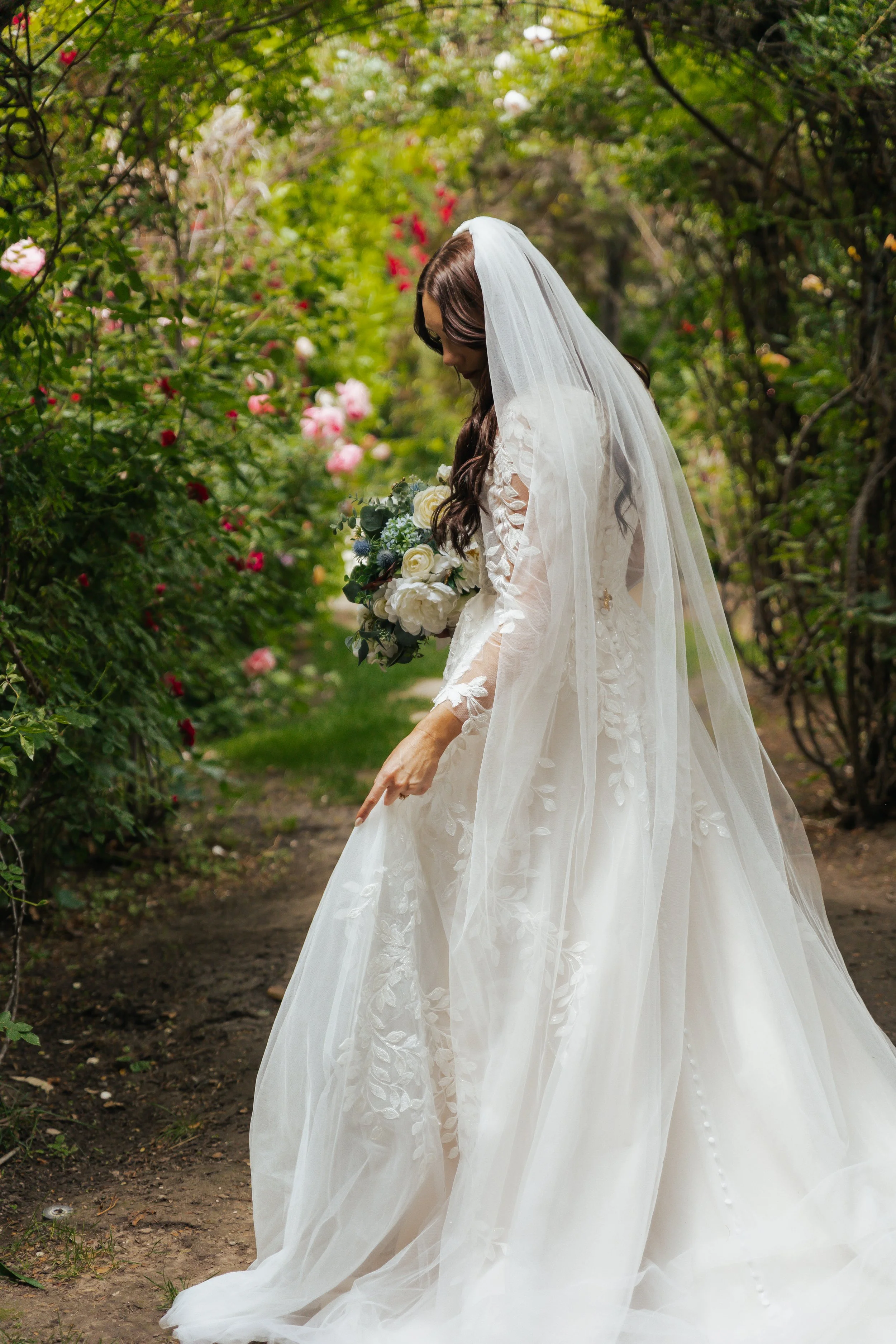 Bride in a white wedding gown holding a bouquet of roses in a garden aisle.