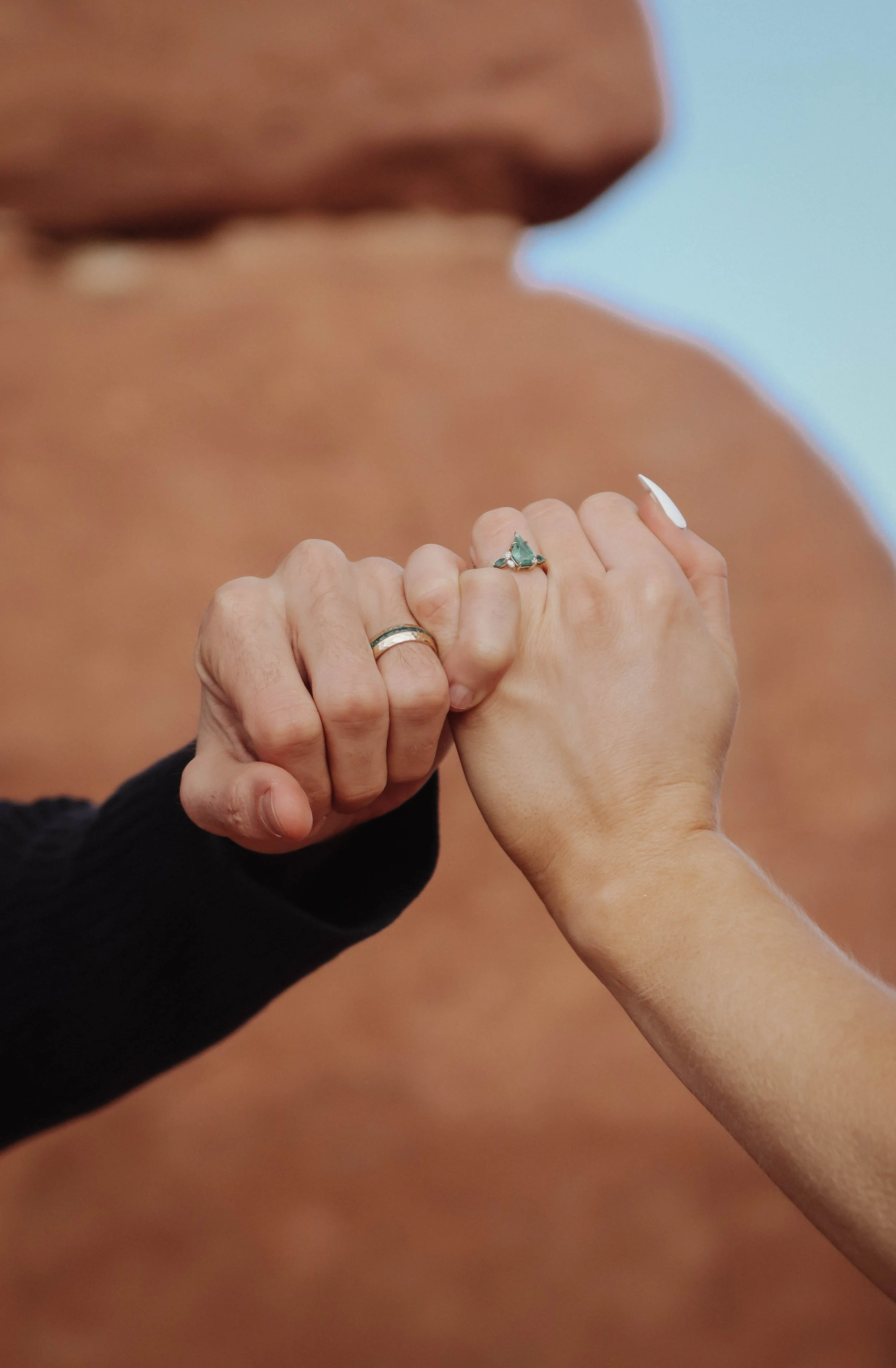 Close-up of two hands touching, each wearing a ring, with a blurred background of a person's face and shoulder.