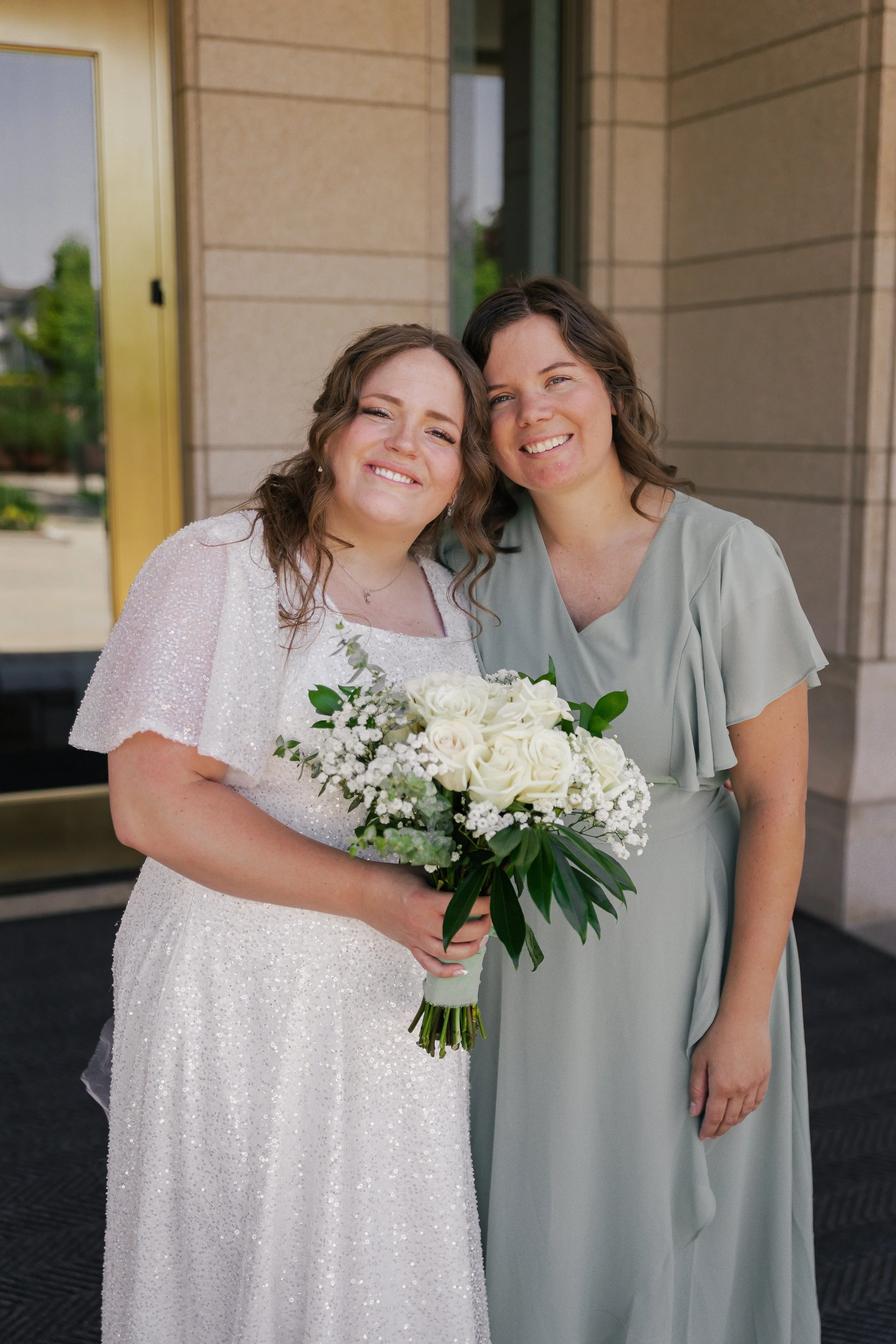 A bride holding a bouquet of white roses and greenery, standing next to a woman in a light green dress, both smiling outside a building.