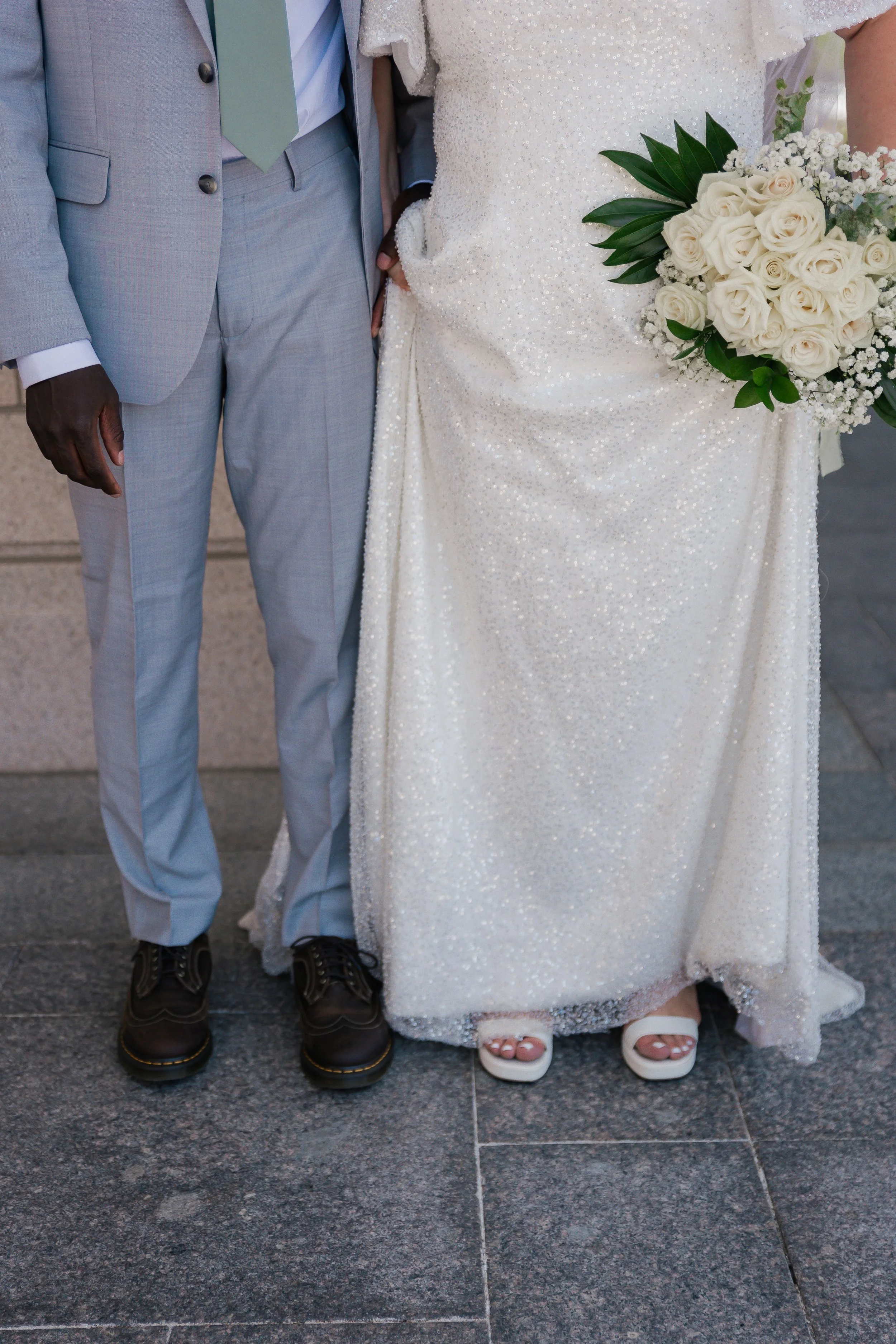 Close-up of a couple at a wedding, showing only their lower bodies. The groom is wearing a light gray suit with black shoes, and the bride is in a white, sparkly wedding gown holding a bouquet of white roses and greenery.