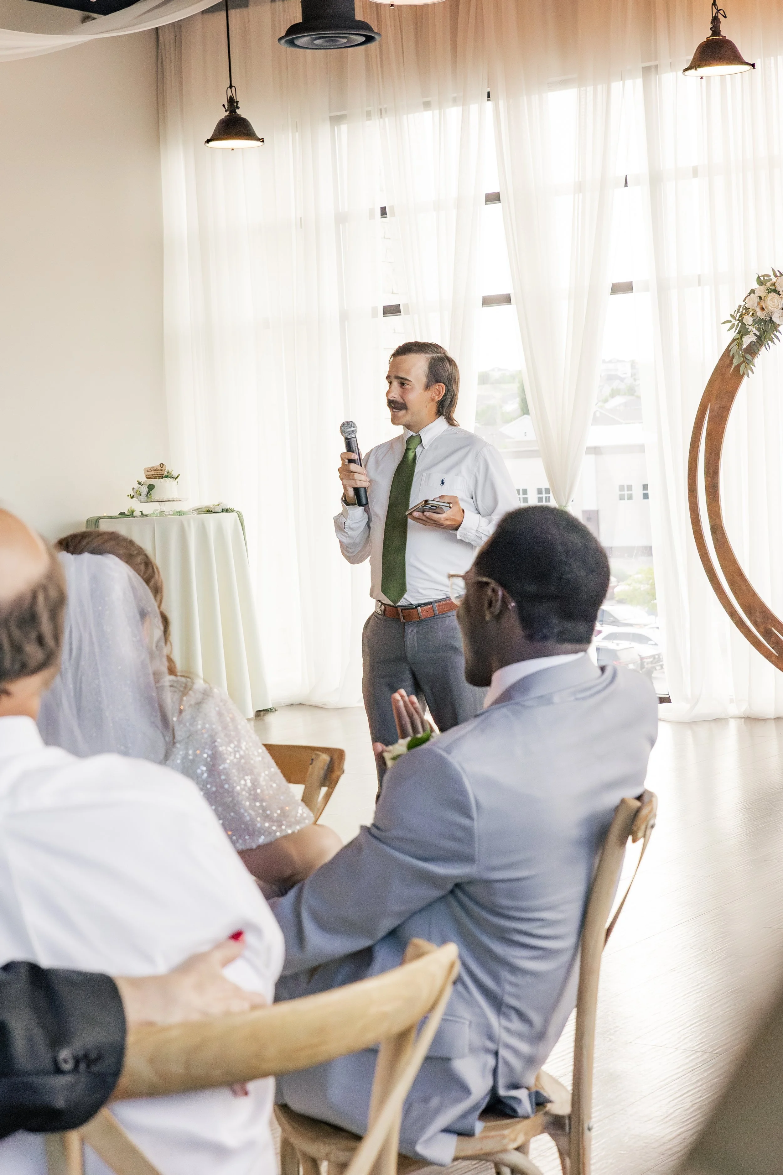 A man with a microphone giving a speech at a wedding reception, with guests seated and listening in a bright room with sheer curtains and ceiling lights.