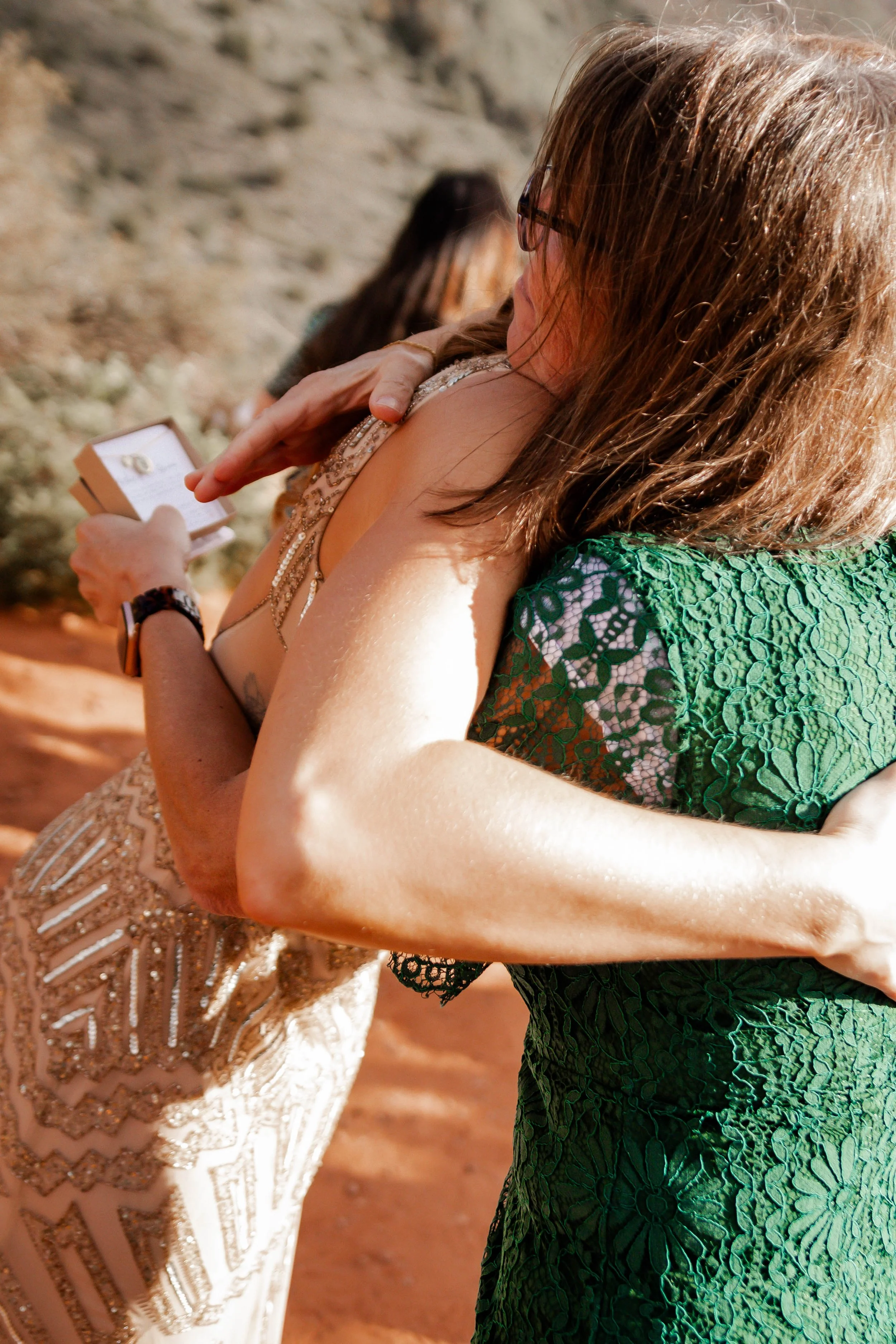 Two women hugging at an outdoor event. One woman is holding a small box, possibly an engagement ring, and appears to be emotional.