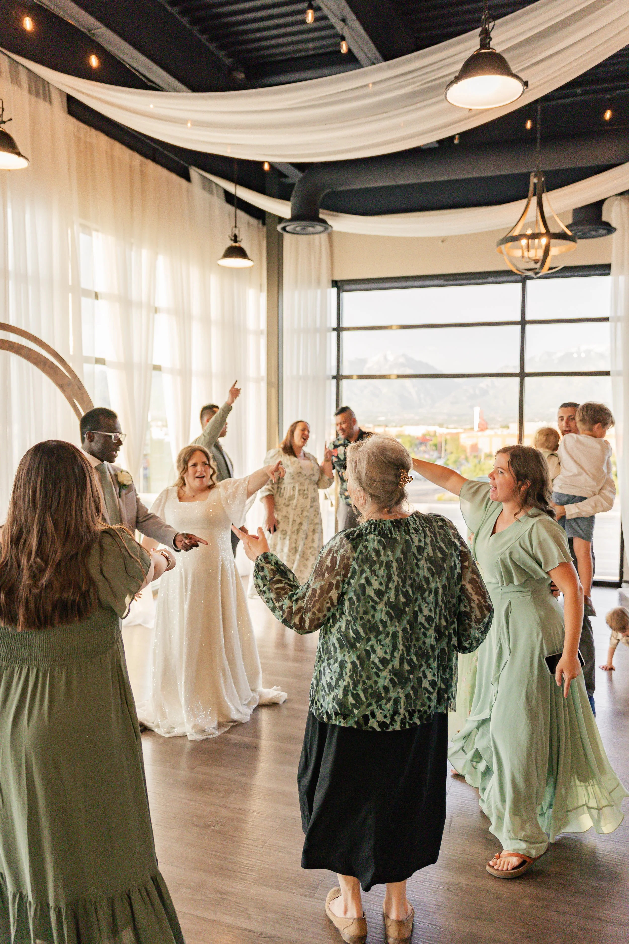 Happy wedding reception with guests dancing and celebrating indoors, featuring large windows with mountain view, and decorated ceiling with white curtains and hanging lights.