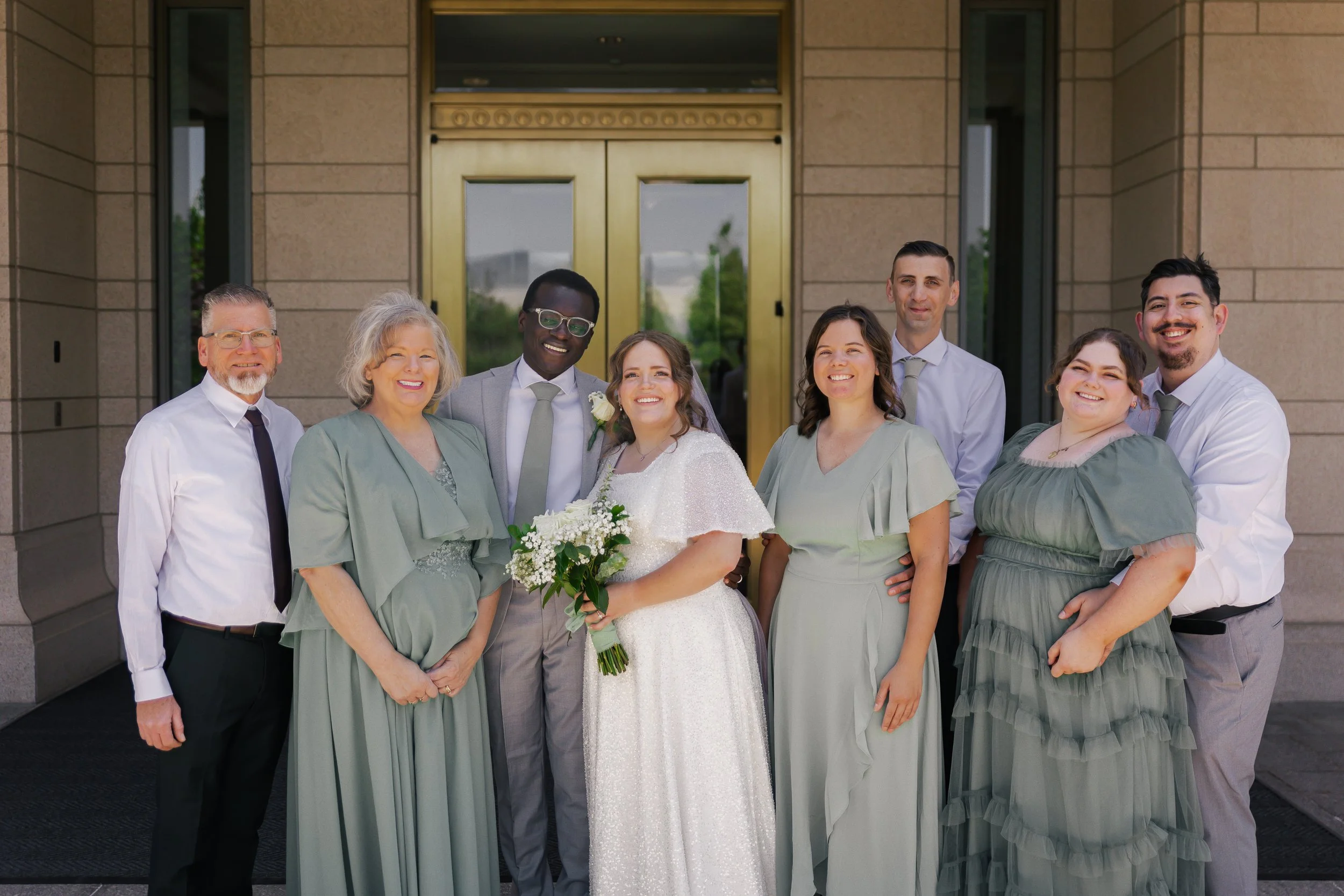 Group of eight people standing outside a building, smiling for a photo. One woman in a white wedding dress holding a bouquet is in the center, surrounded by friends and family dressed in formal attire.