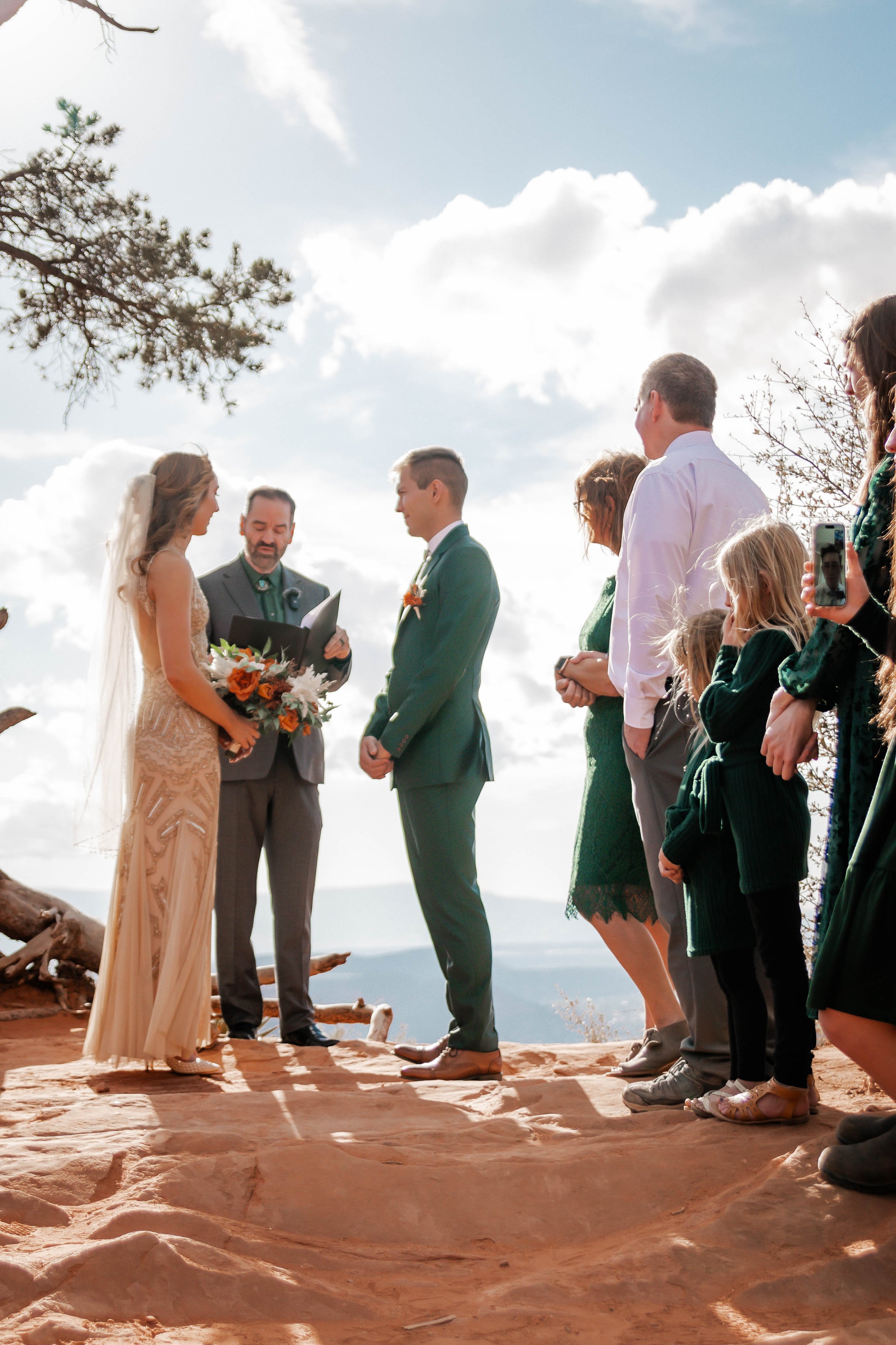 A couple getting married during an outdoor ceremony with the officiant and guests standing on a rocky surface under partly cloudy sky.