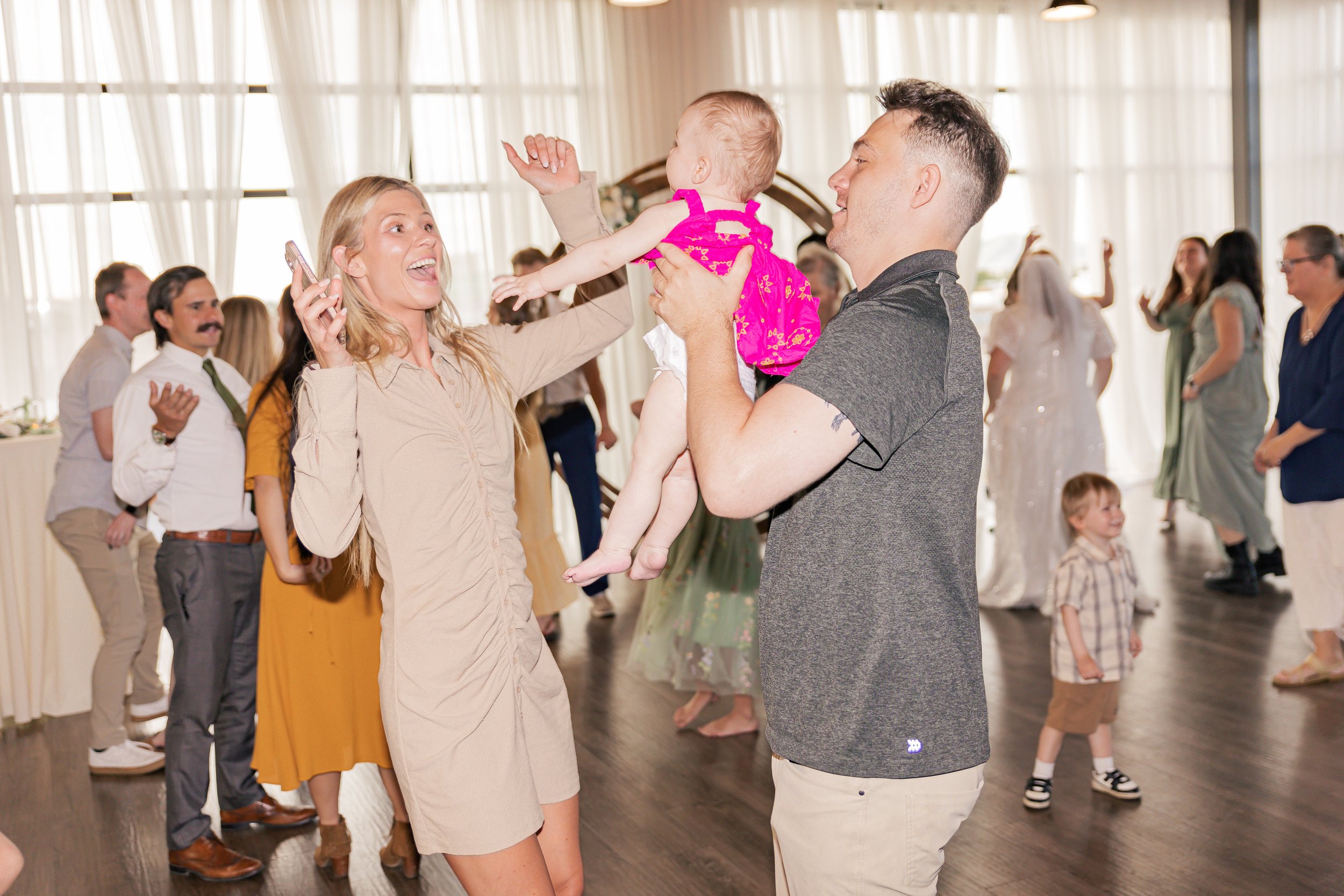 A woman and a man at a celebration, with the woman holding a phone and both smiling as they dance. The woman is smiling and raising her arms, the man is holding a young girl dressed in pink, and there's a group of people dancing in the background in 
