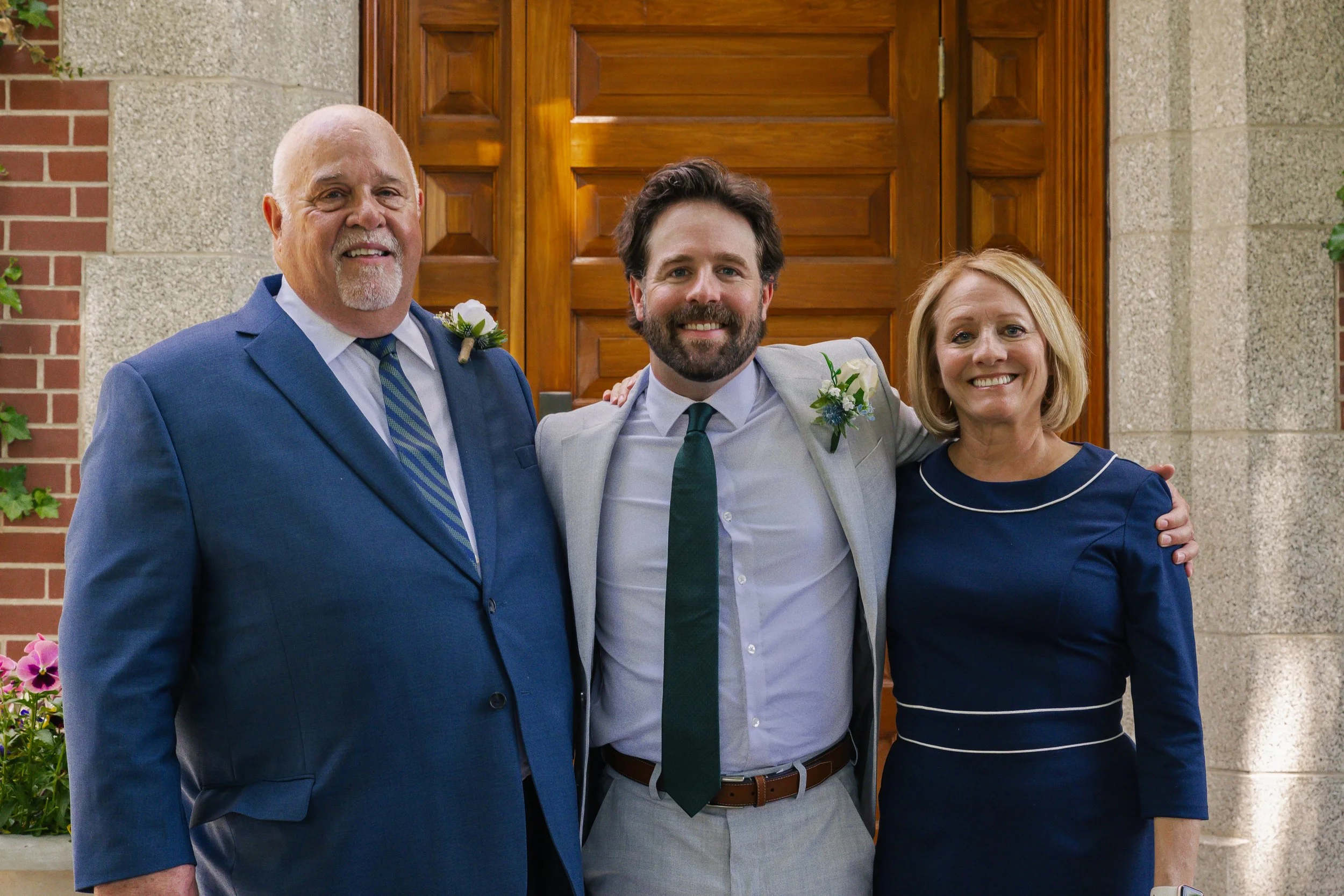 Three people smiling and standing close together in front of a wooden door, dressed in formal attire with white flower boutonnieres, indicating a wedding celebration.