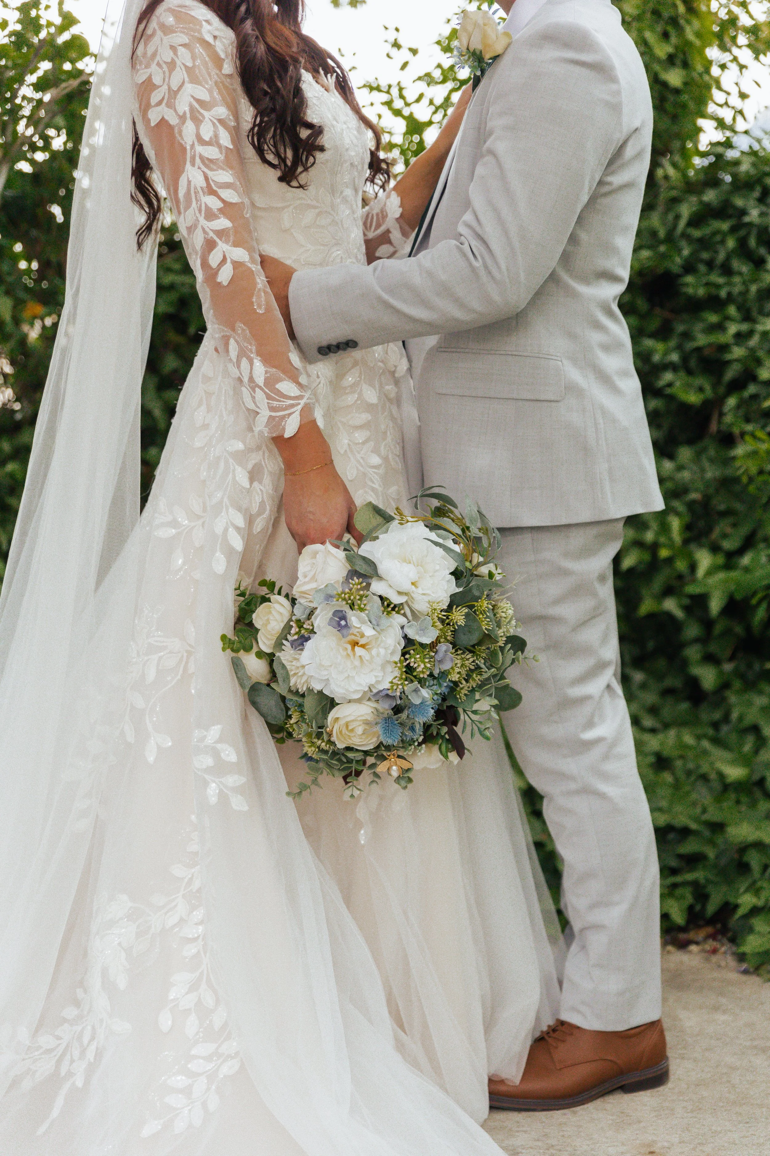 Close-up of a bride and groom holding hands during their wedding ceremony. The bride is wearing a white lace wedding gown with floral embroidery, and the groom is dressed in a light grey suit. The bride is holding a bouquet of white and blue flowers 