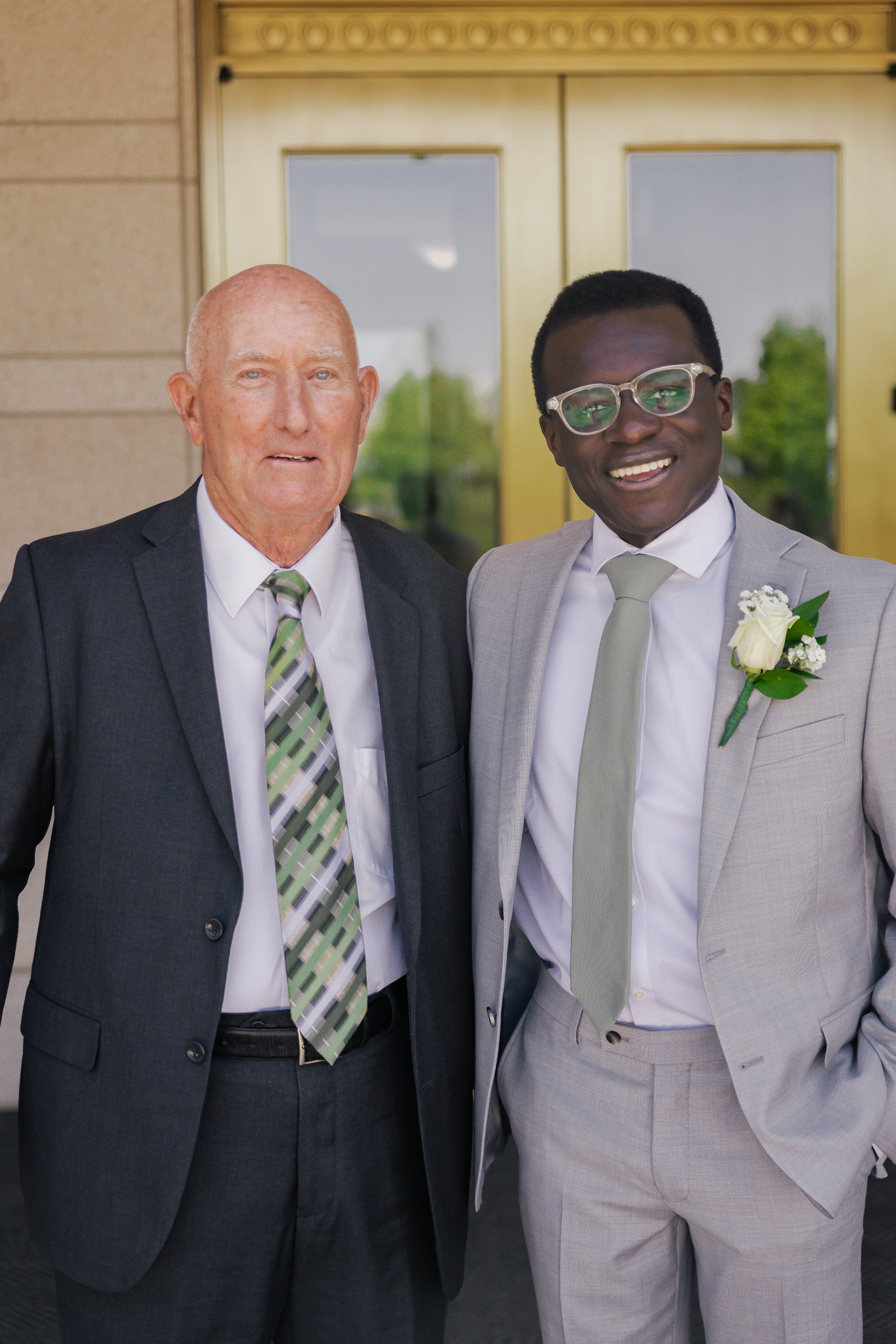Two men in suits standing outside a building, smiling for a photo. One young man wears glasses and a gray suit with a boutonniere, and an older man wears a dark suit with a striped tie.