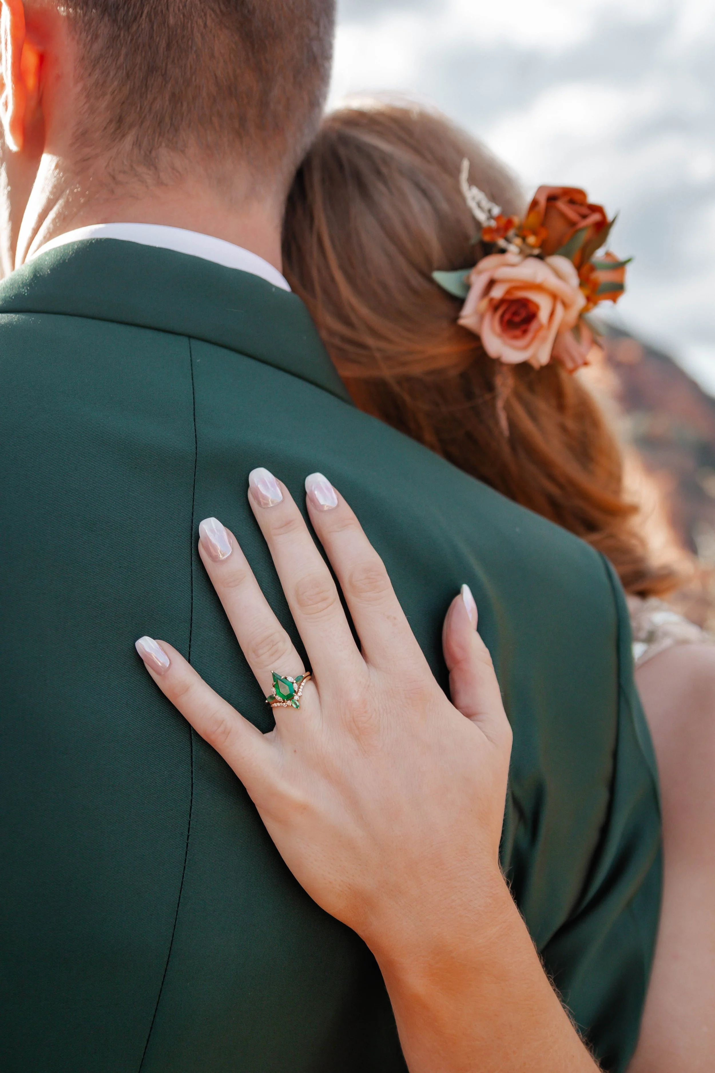 Close-up of a woman’s hand with an emerald and diamond ring resting on a man’s shoulder, both in formal attire, with a flower accessory in the woman’s hair.