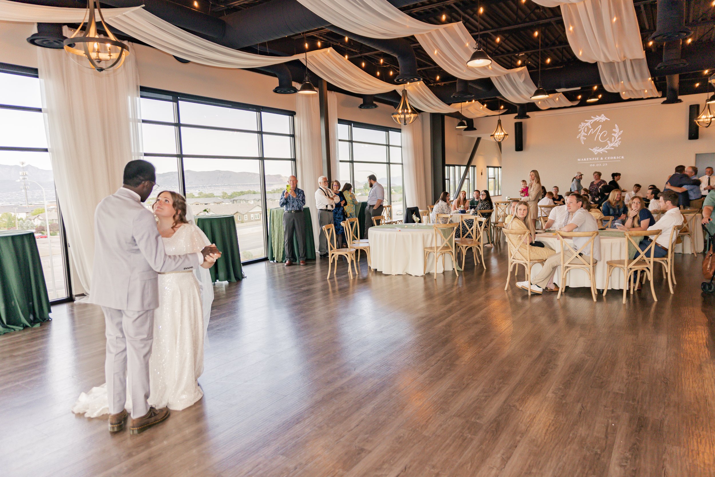 A bride and groom dancing at their wedding reception in a spacious modern event hall with large windows, white drapes, and string lights, while guests sit at round tables and watch.