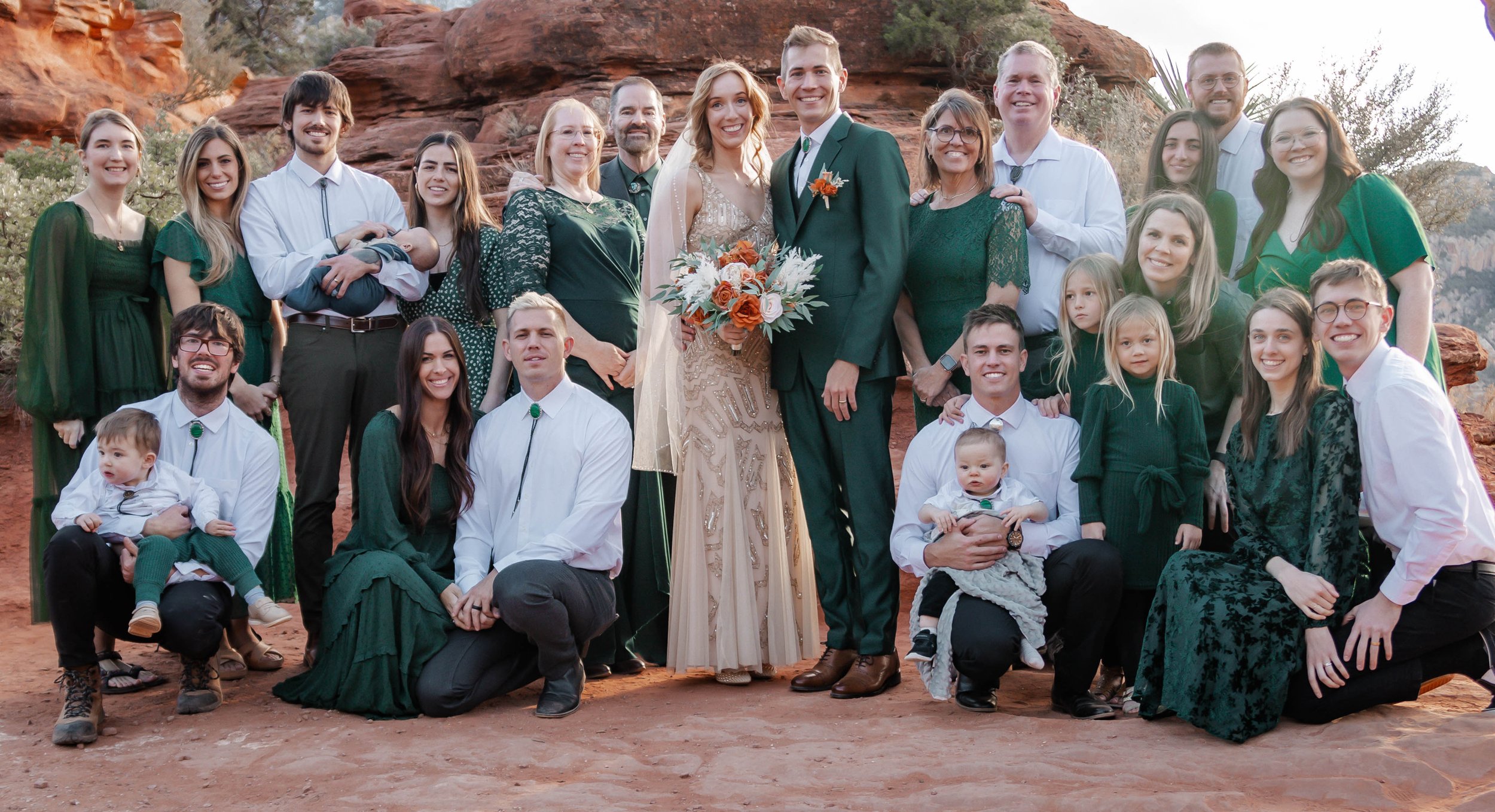 A large group of people gathered outdoors for a wedding photo, standing on reddish rock with red cliffs and desert vegetation in the background. The bride is holding a bouquet and the groom is wearing a green suit. The group consists of men, women, a