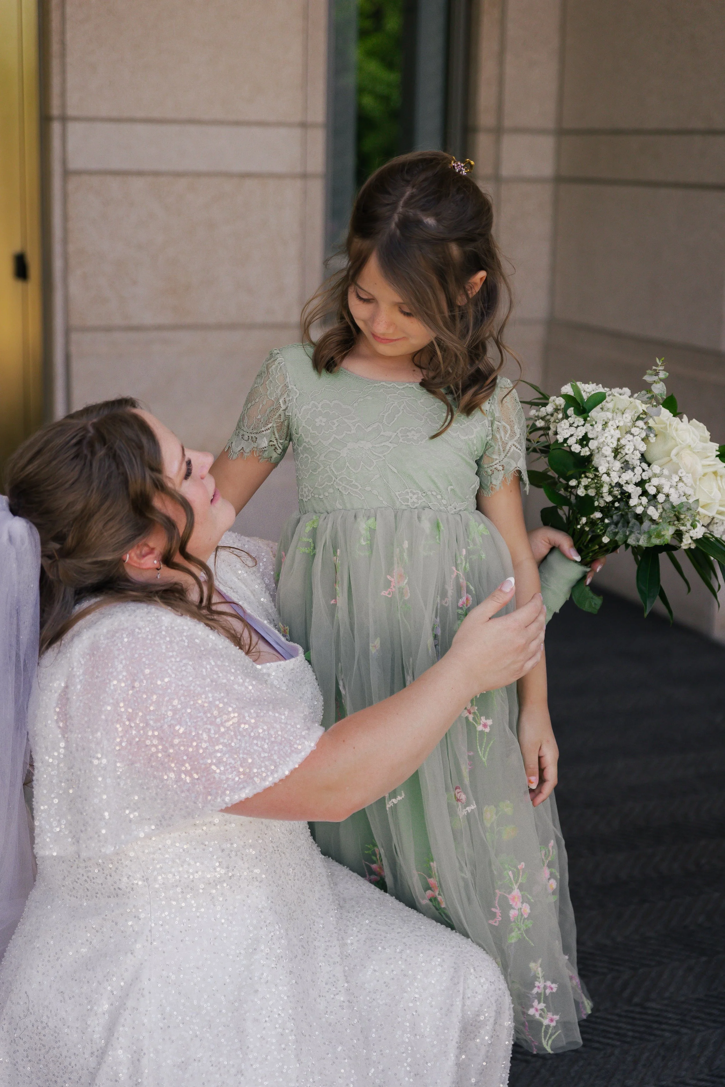 A woman in a sparkly white dress is sitting and looking up at a young girl in a light green dress holding a bouquet of white flowers, both sharing a tender moment.