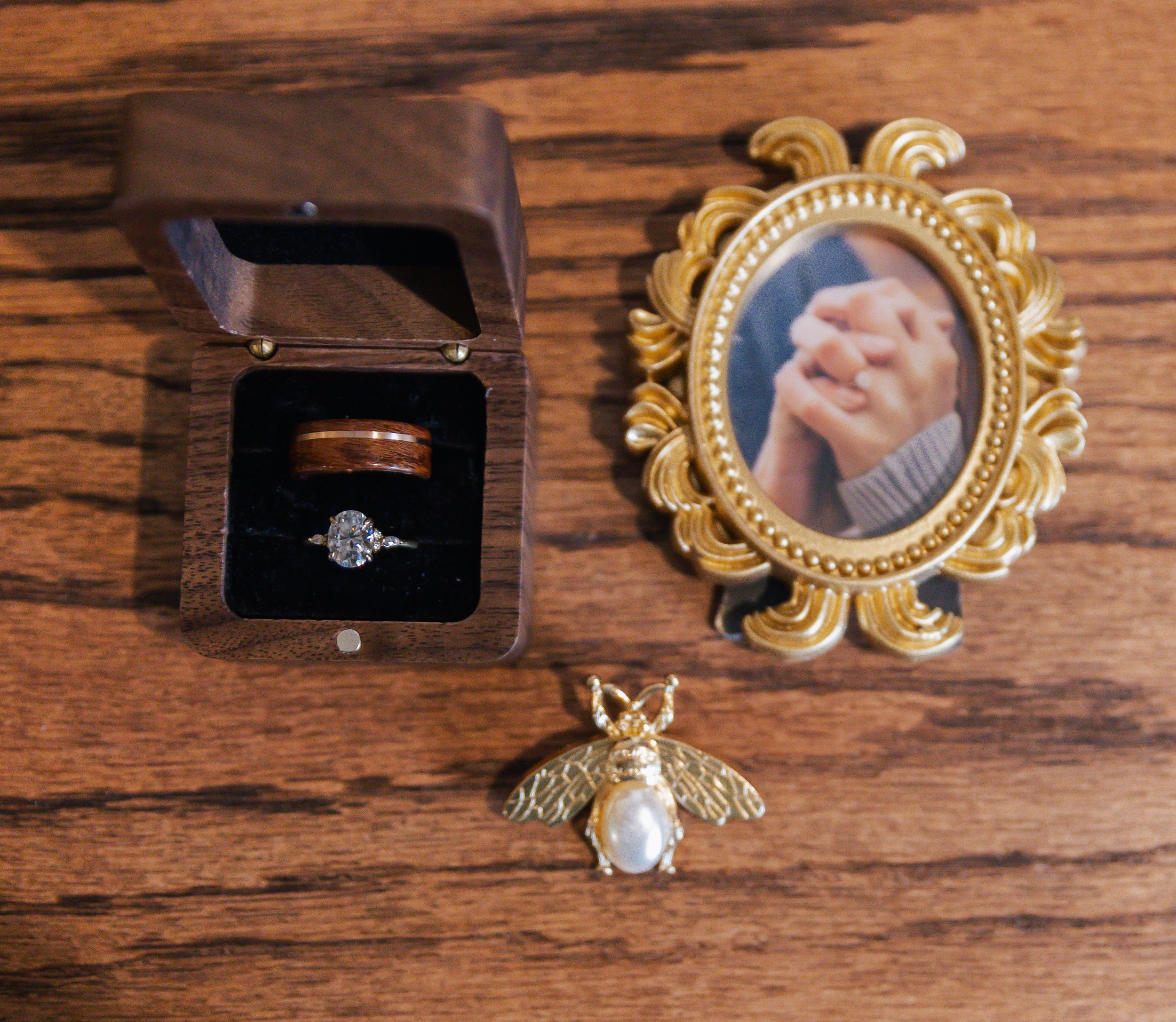 A wooden ring box with a diamond engagement ring inside, a gold-framed photo of hands holding, and a gold bee pin with a pearl body on a wooden surface.
