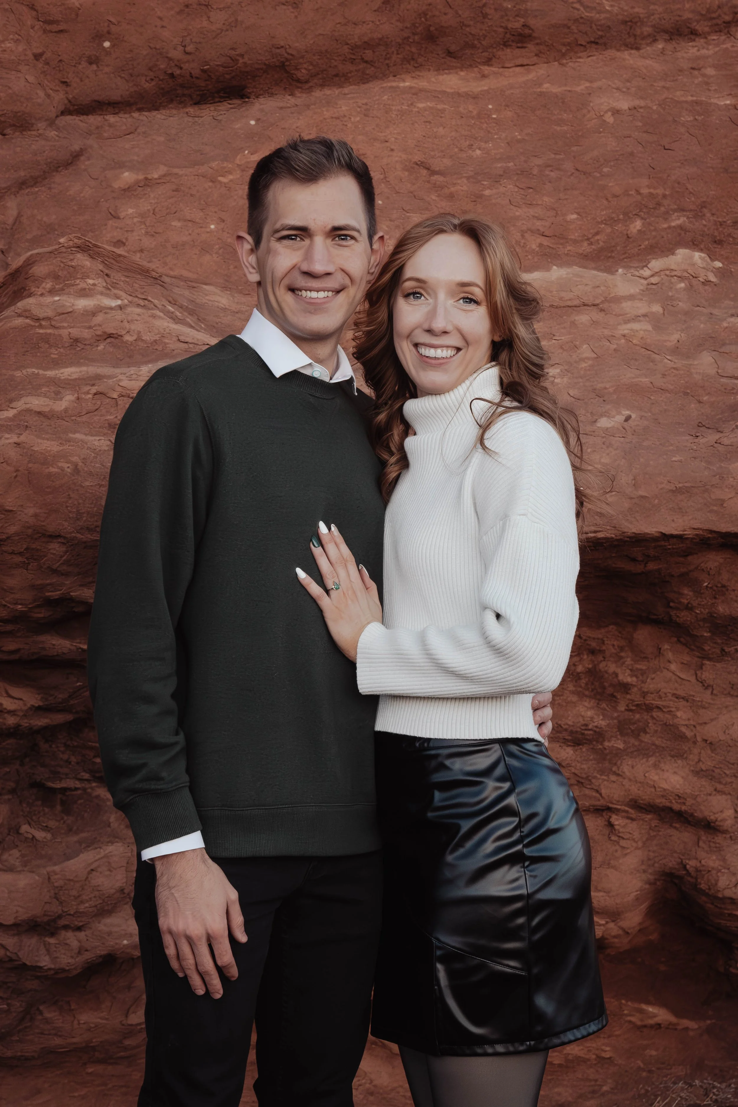 A smiling man and woman standing together against a red rock background, with the woman's hand on the man's chest displaying a ring.