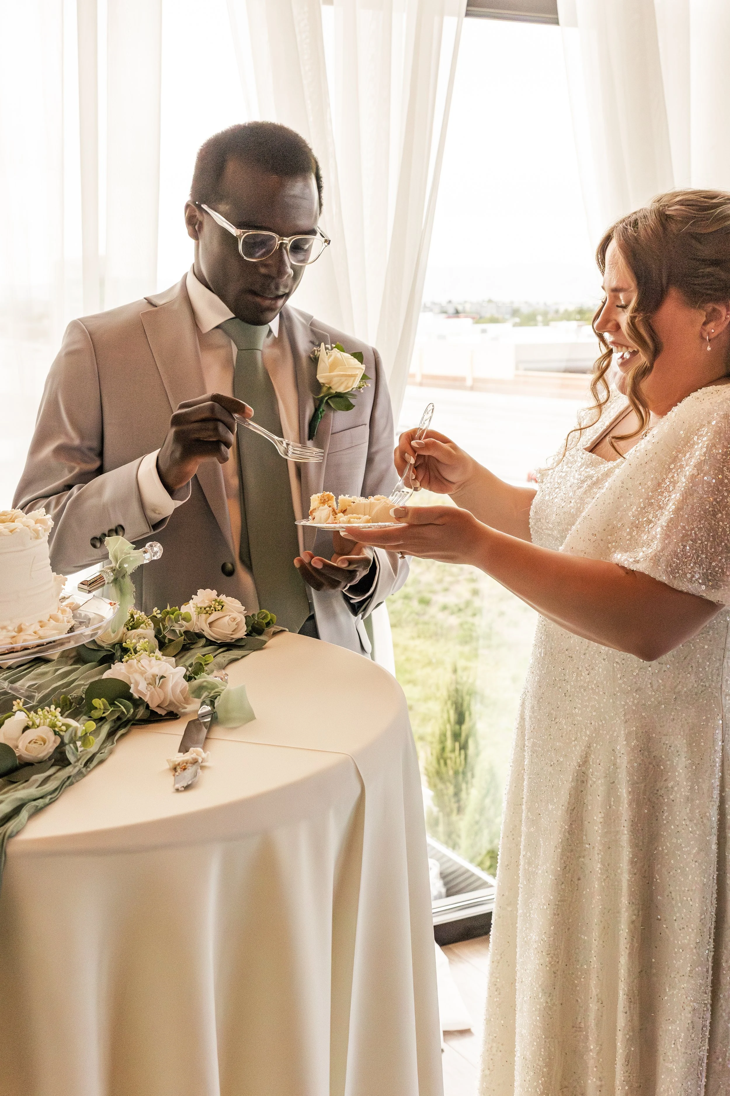 A bride and groom at their wedding reception sharing a piece of cake. The groom is dressed in a light gray suit with a green tie and boutonniere, while the bride is in a white, sparkly dress. They are smiling and holding cake plates with utensils. Th
