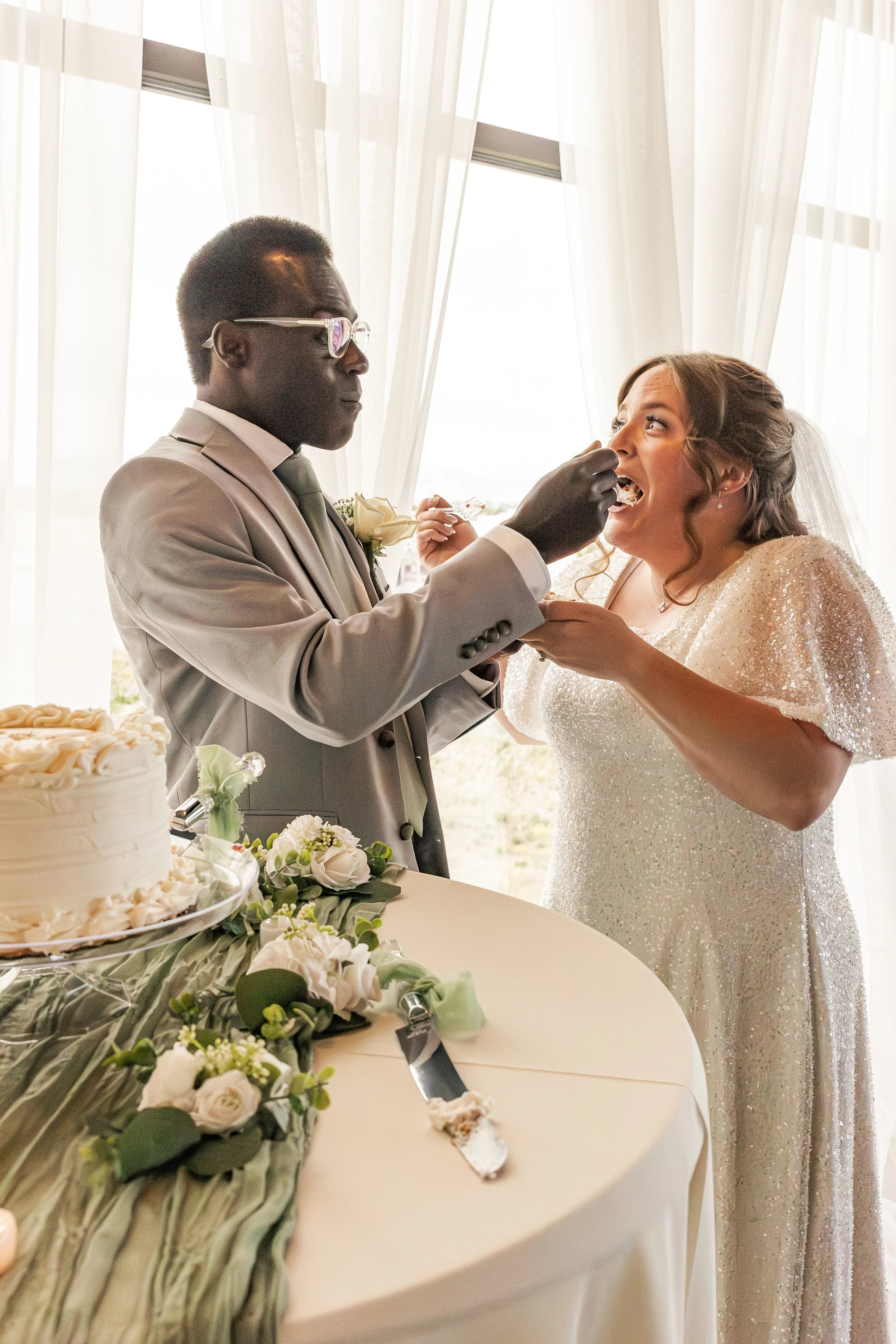 A bride and groom at their wedding reception, with the groom feeding the bride a piece of cake, standing near a table with wedding cake and floral arrangements.